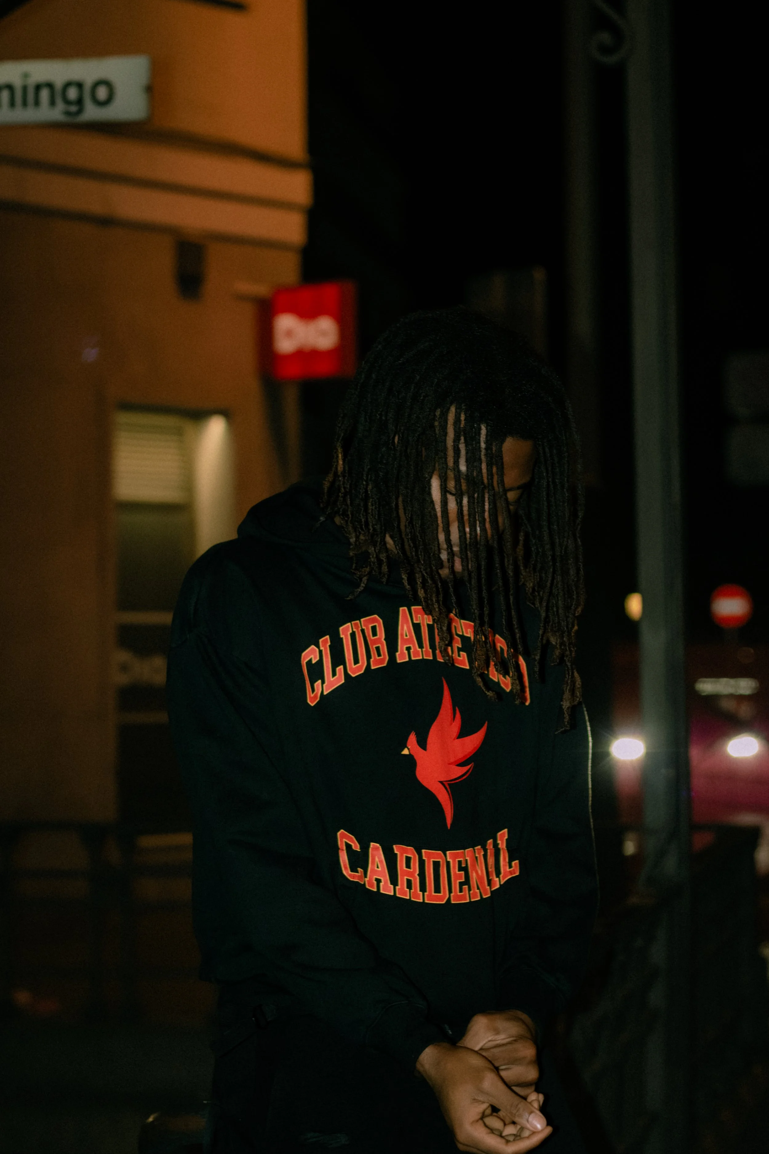 A person with long braided hair wearing glasses and a dark hoodie with 'CLUB AT CEDAR Carden' text and a red phoenix logo, standing in a dimly lit area at night.