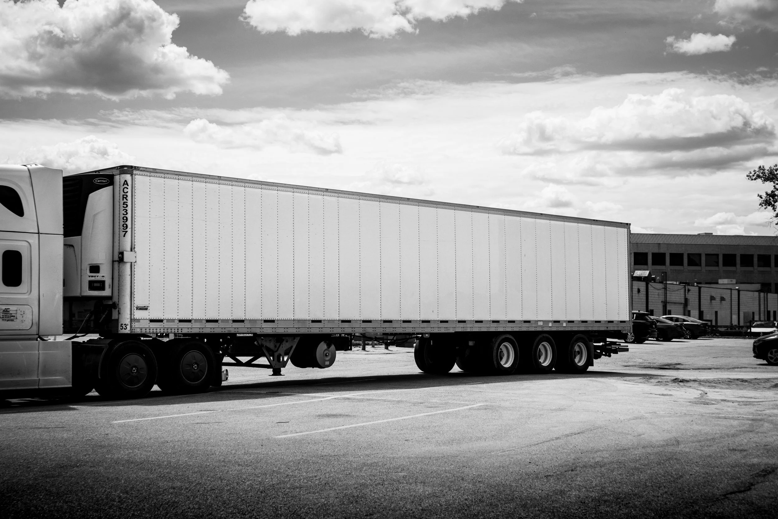 Black and white photo of a semi-truck parked in an outdoor lot with a building and cars in the background, under a cloudy sky.