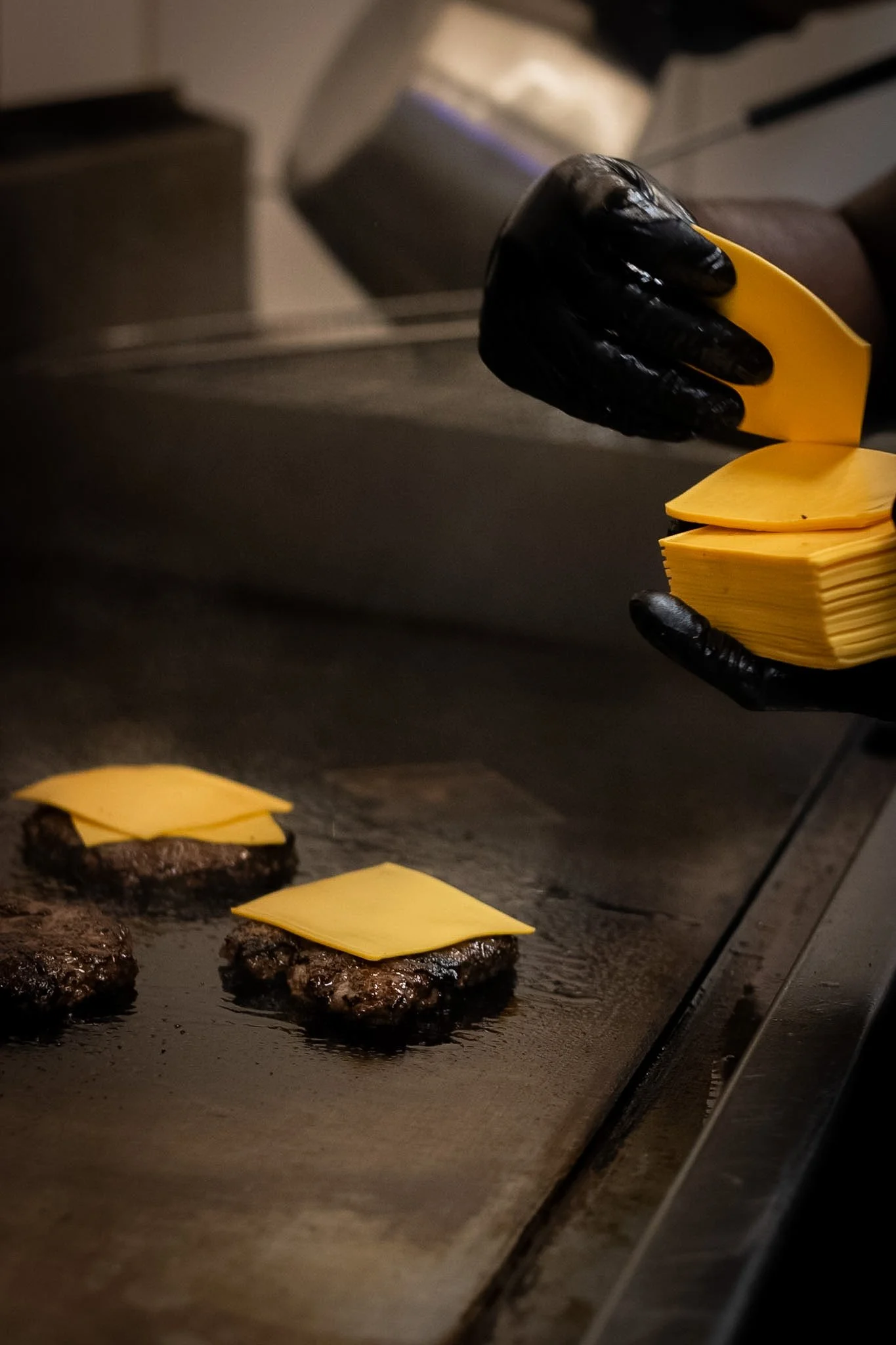 Close-up of burger patties with slices of cheese on a flat-top grill, with a hand placing or removing a slice of cheese using a yellow spatula.
