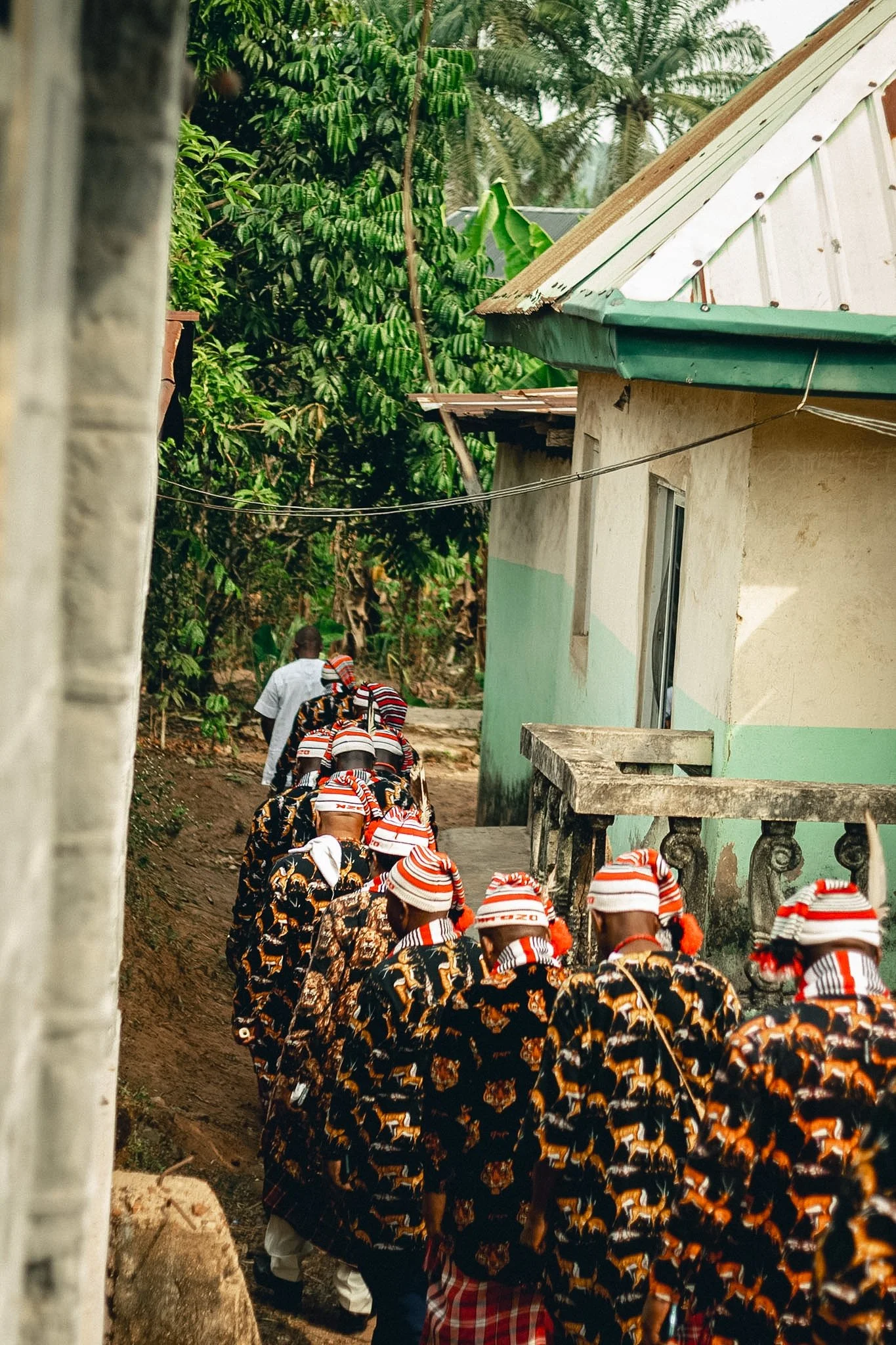 A group of children at a funeral procession in a rural village, wearing matching outfits with orange hats and patterned dresses, walking in a line past a house with green and white walls, surrounded by trees and greenery.