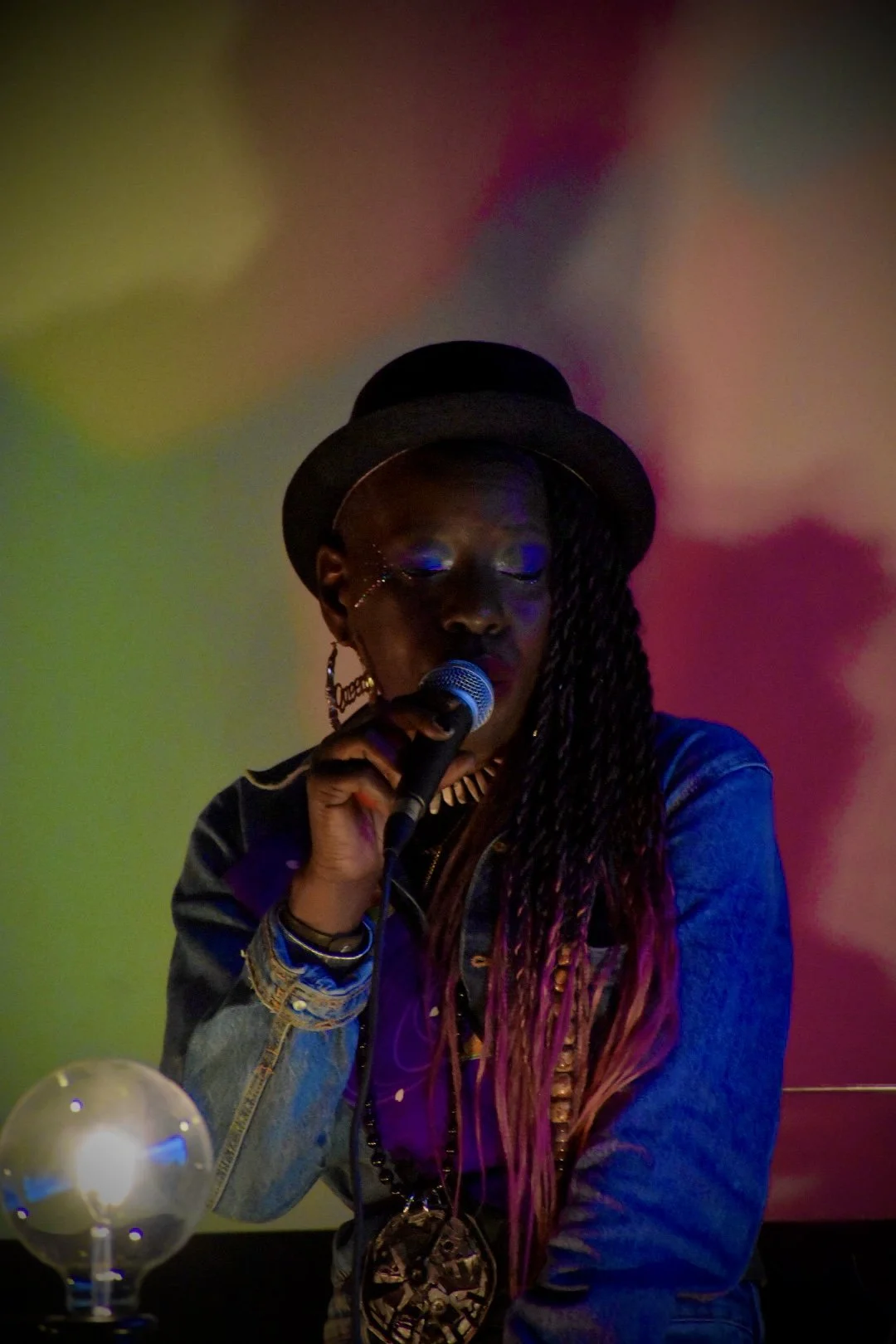 A woman with dreadlocks wearing a black hat, large hoop earrings, and a denim jacket, holding a microphone and performing or speaking in front of a colorful abstract background.