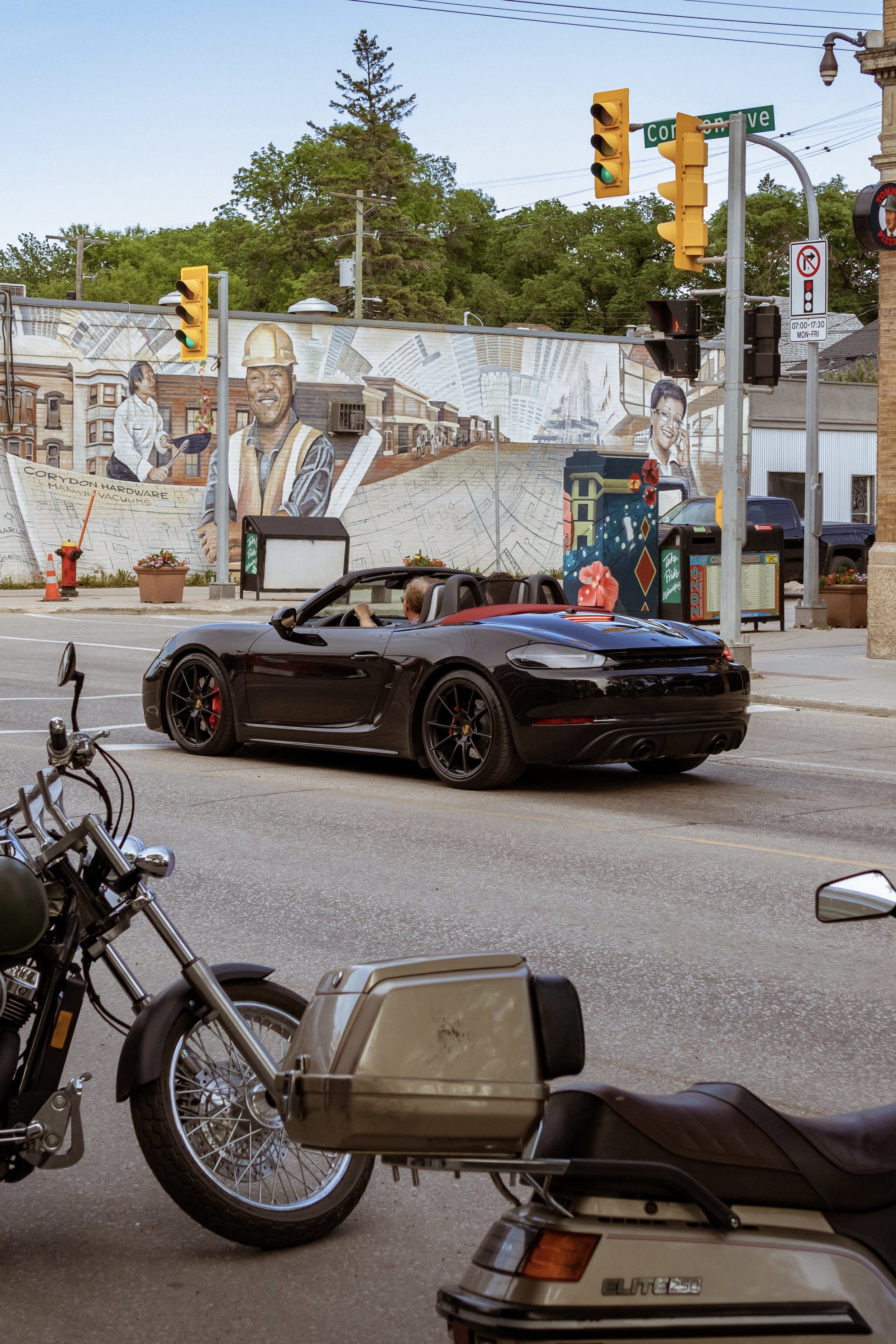A city street intersection with traffic lights, parked motorcycles, a black sports car, a black pickup truck, and a mural on a building wall depicting construction workers, buildings, and urban scenes. Street signs and a trash receptacle are also vis