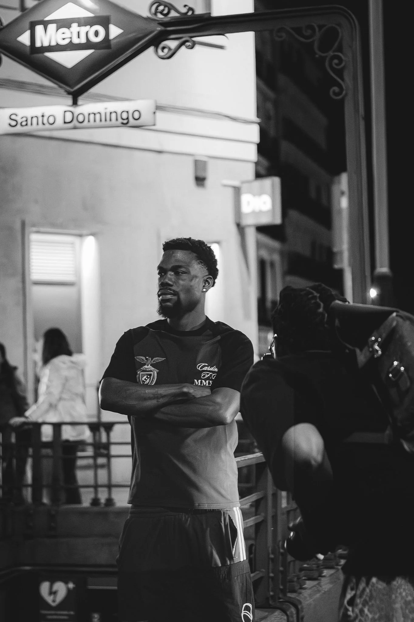 A man with short curly hair and beard standing with crossed arms in an outdoor setting at night. Behind him, a woman with long hair is talking to someone out of frame. There are street signs, including a Metro sign and a street sign for Santo Domingo