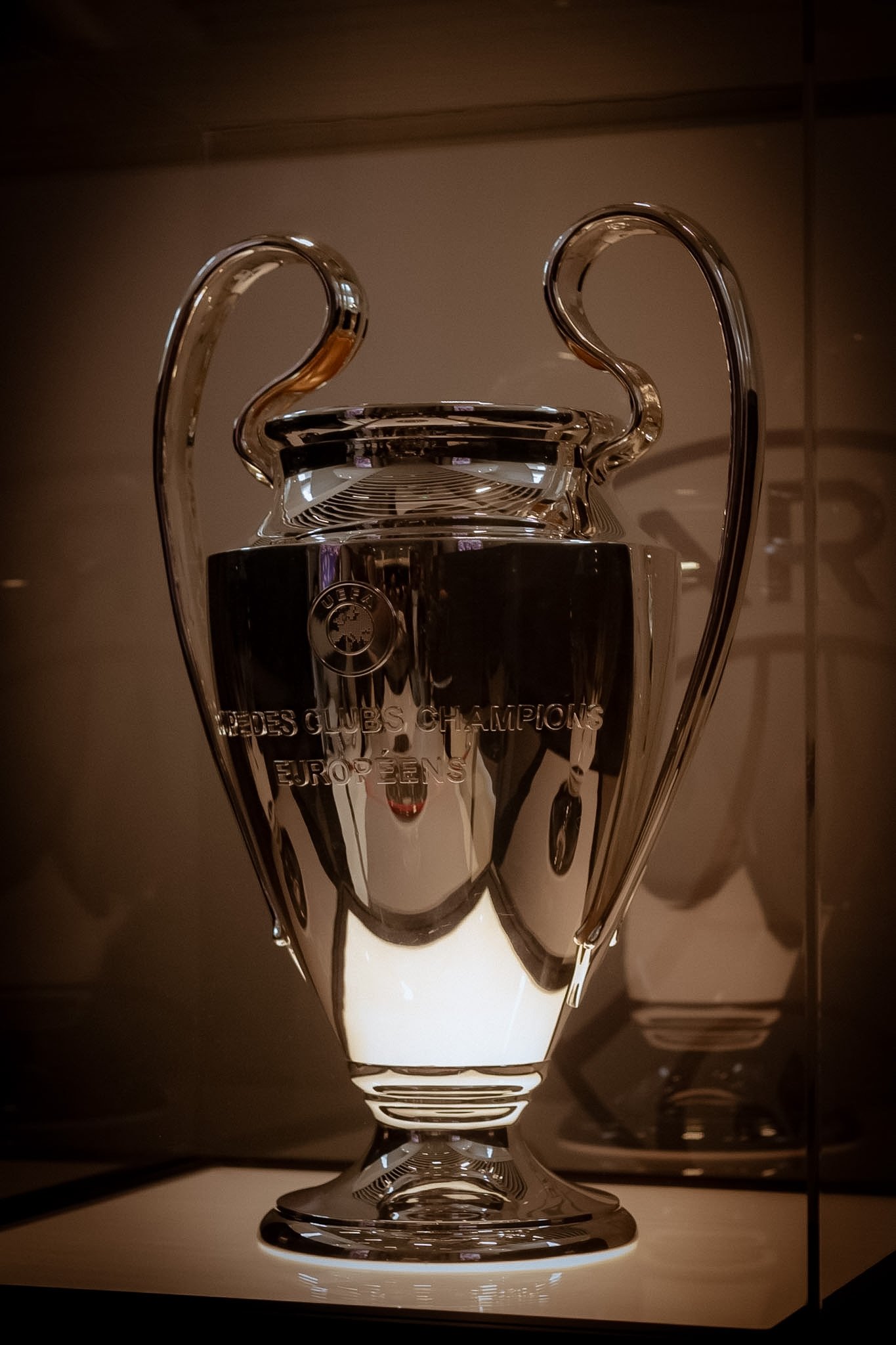 A glass trophy with a sleek design, engraved with 'UEFA CLUBS CHAMPIONS' and 'EUROPE'. The trophy is illuminated from below, casting shadows on the surface.