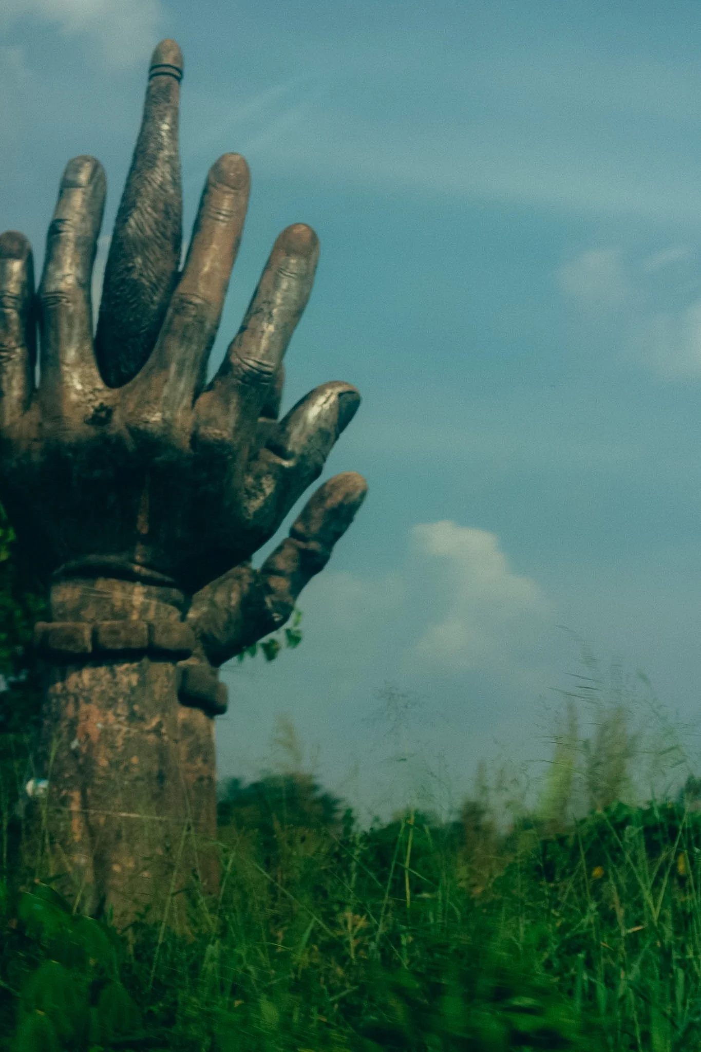 A bronze-colored statue of a hand with extended fingers, placed on a stone pedestal outdoors, with green grass and trees in the background under a cloudy sky.