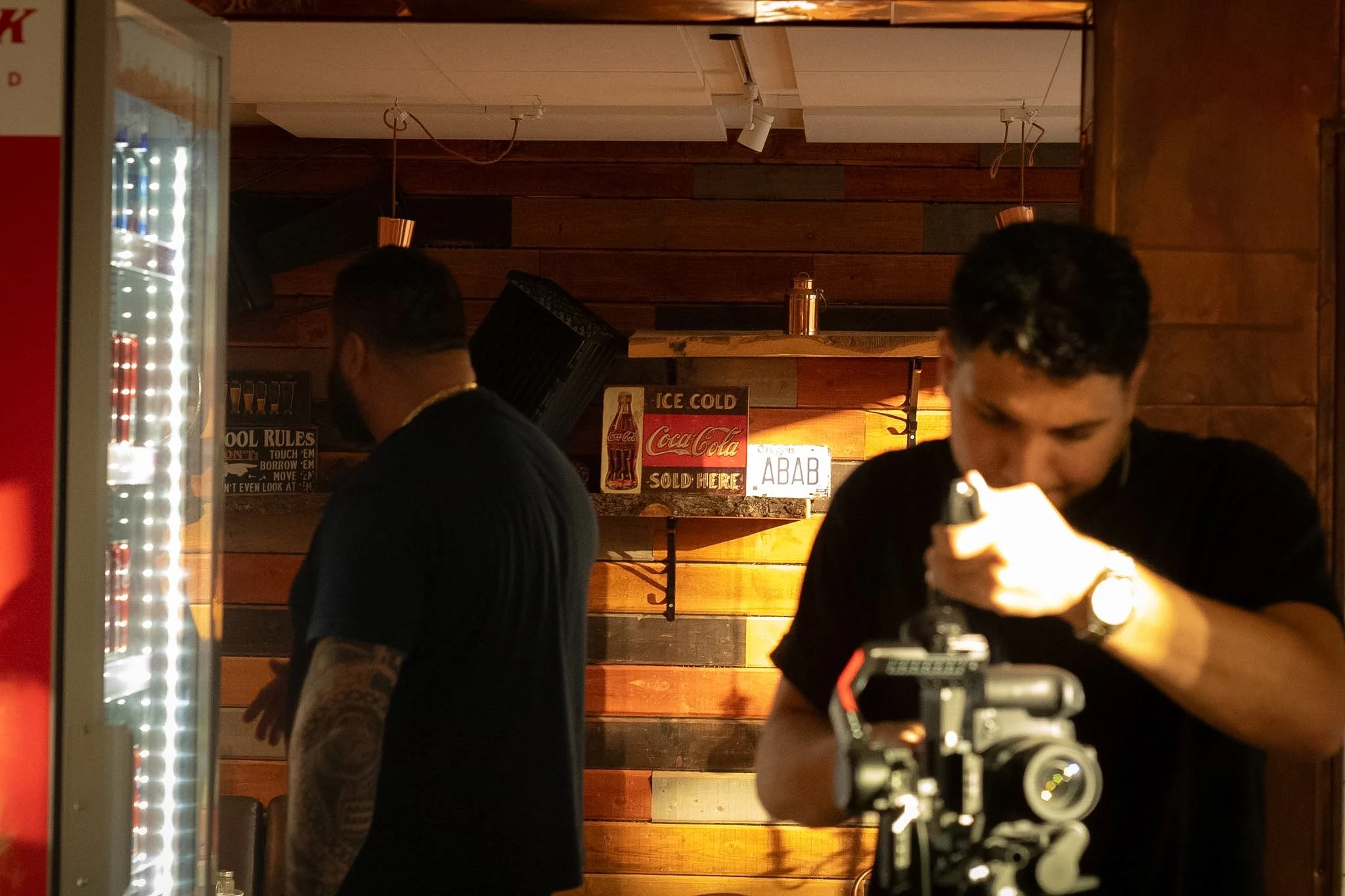 A man in a black shirt is looking at a camera, and two other men are facing away, inside a room with wooden walls.