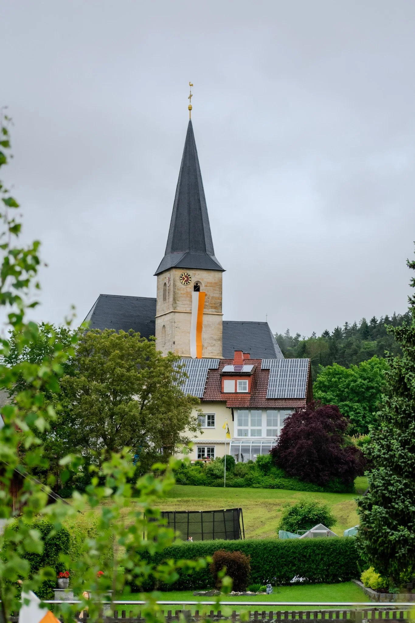 A church with a tall steeple topped with a weather vane, surrounded by lush green trees and a garden with a fountain, and a building with solar panels on its roof.