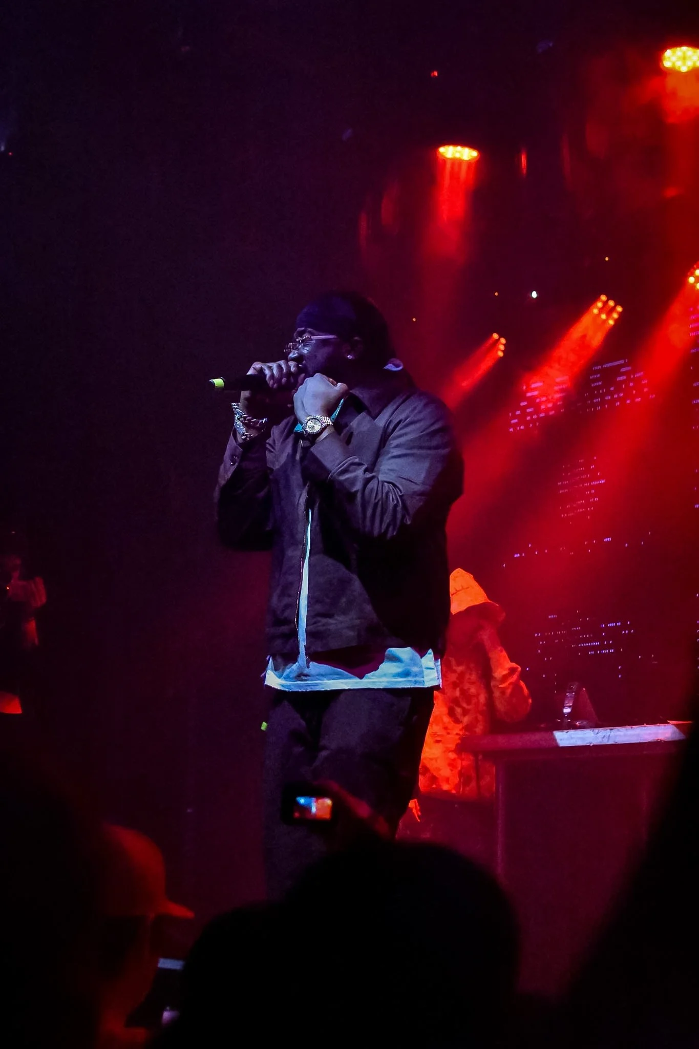 A performer with glasses and a jacket holding a microphone on stage under red and orange lighting, with an audience taking photos and a person in an orange hat visible in the background.