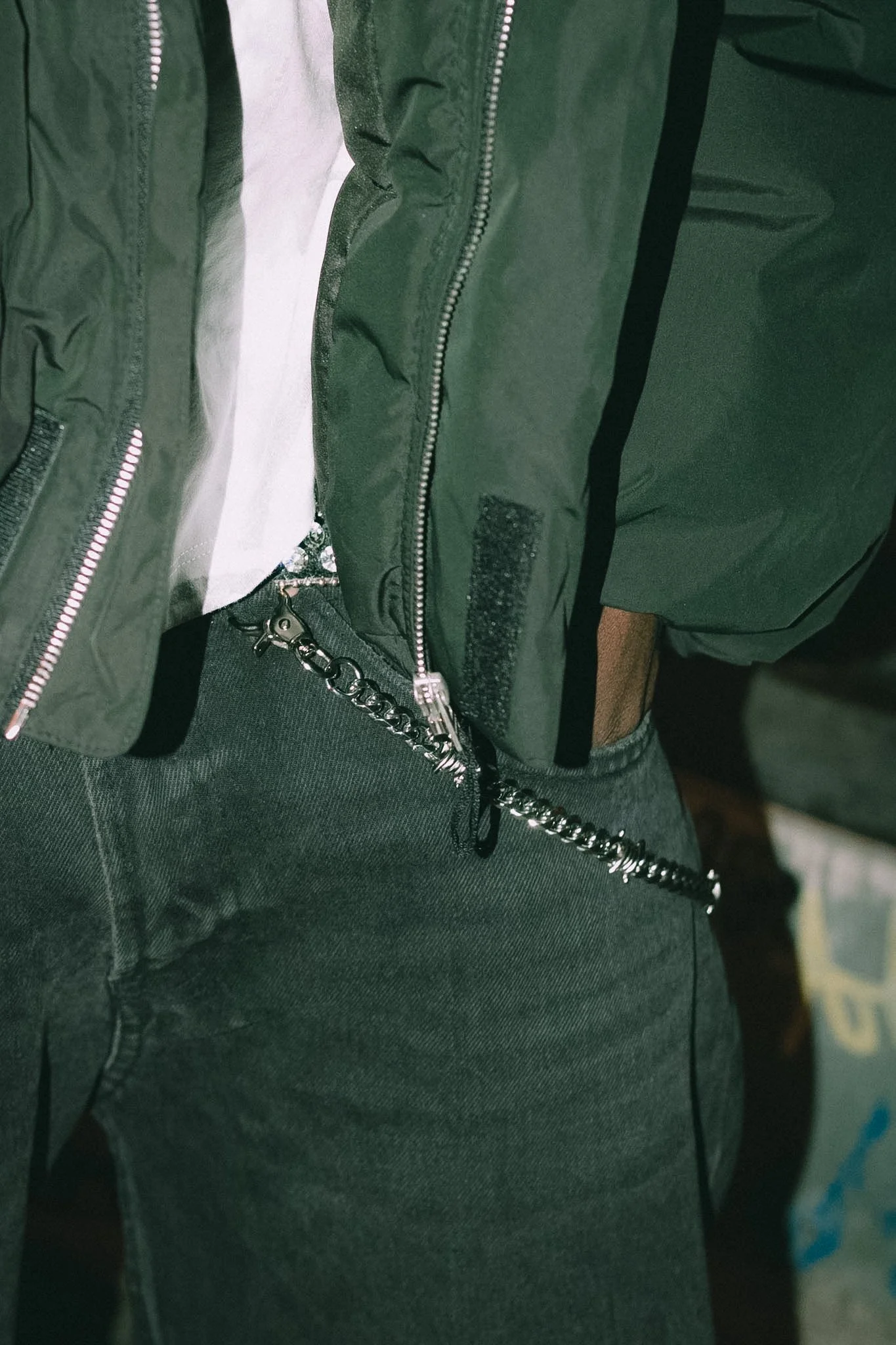 Close-up of a person's waist showing a black chain attached to their dark gray pants with a zipper. The person is wearing a green jacket with a black stripe.