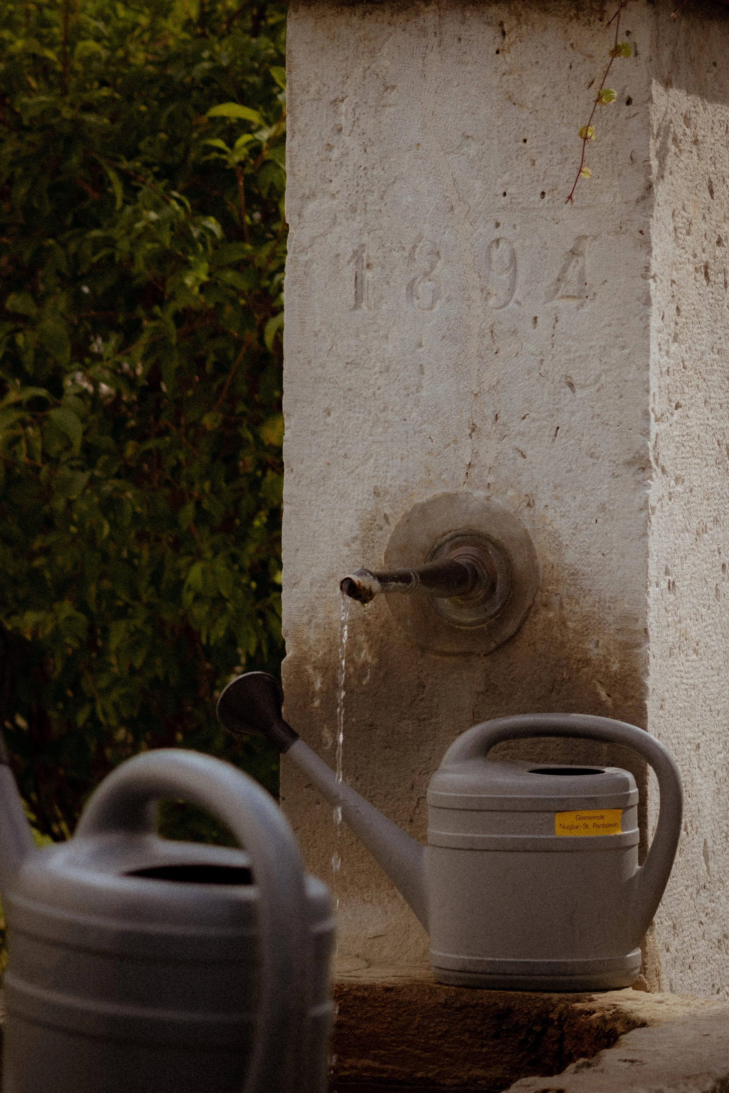 A stone water fountain with a spout dripping water, surrounded by grey watering cans and a white container with a yellow label, outside with greenery in the background.