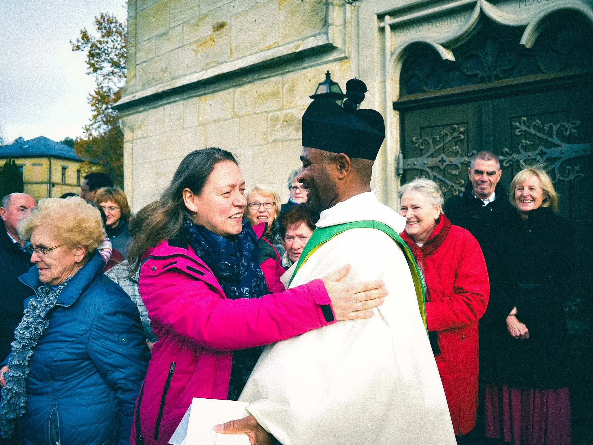 A group of people gathered outside, with a man in a white robe and black hat greeting a woman in a pink jacket by touching her hand, in front of a stone building with ornate metal doors.