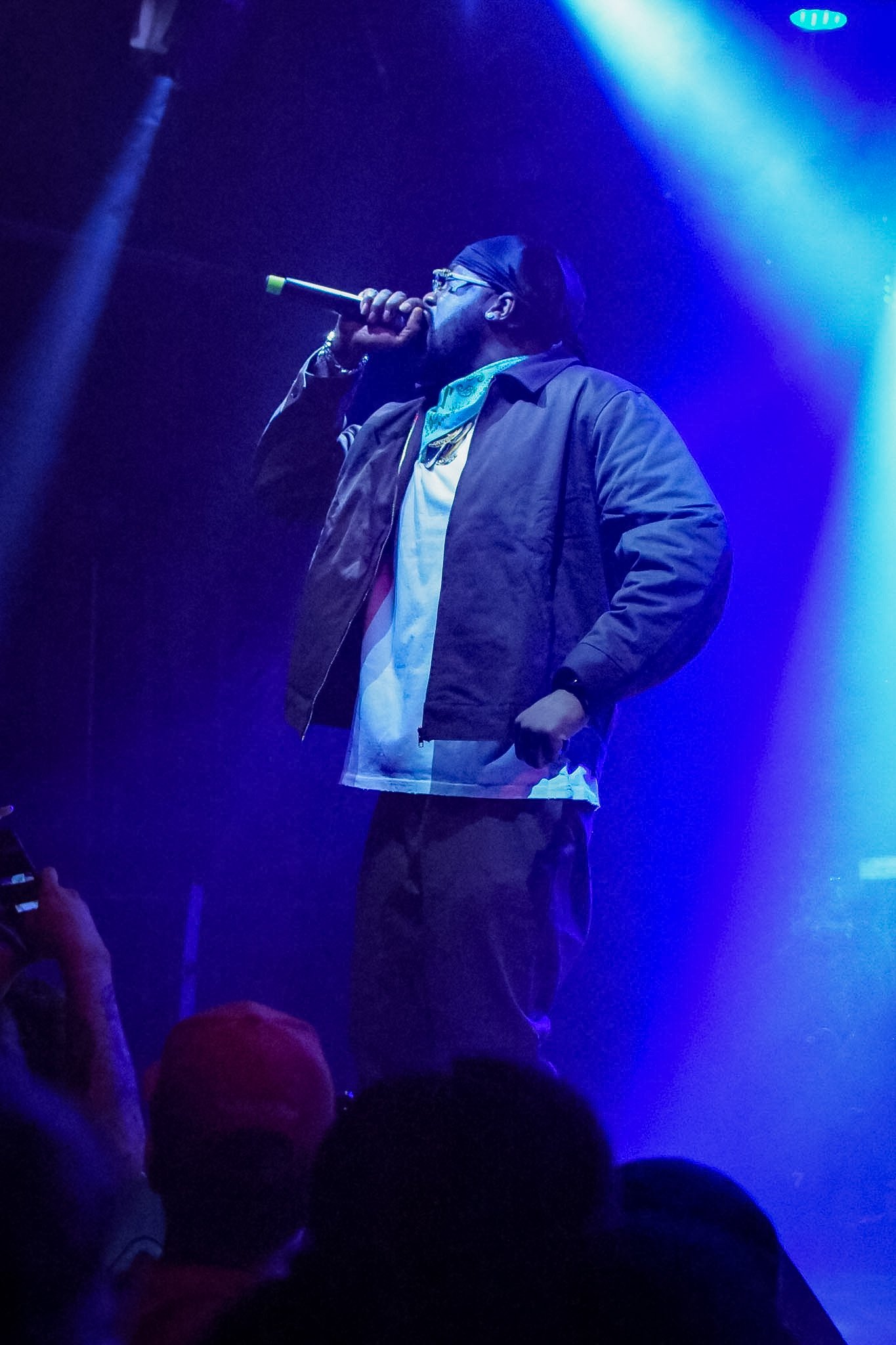 A man wearing glasses, a cap, and a jacket is holding a microphone on stage, illuminated by blue stage lighting, with an audience visible in the foreground.