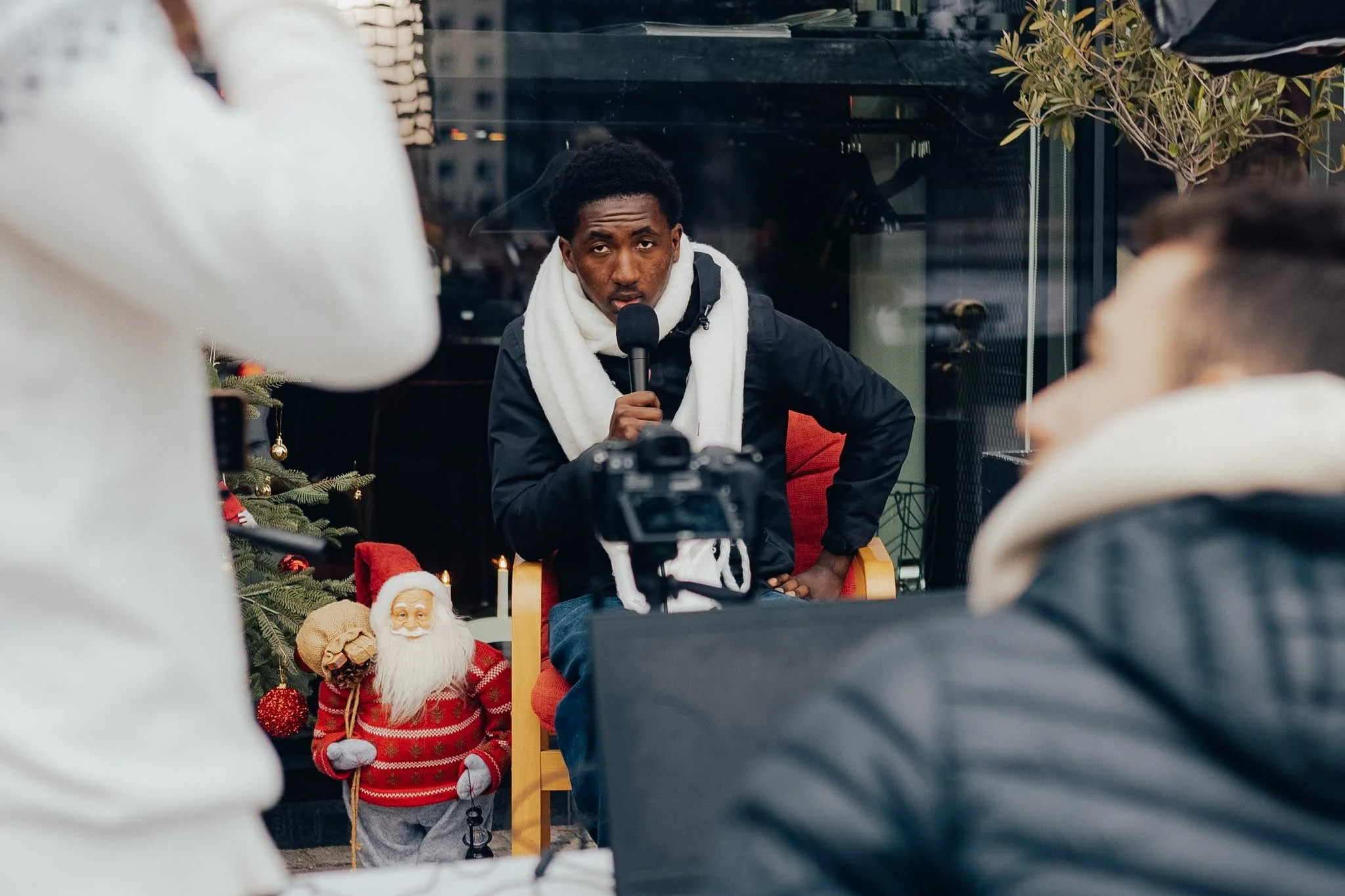 A man sitting indoors holding a microphone, wearing a white towel around his neck, with Christmas decorations including a decorated tree and Santa Claus figure nearby, and people around.
