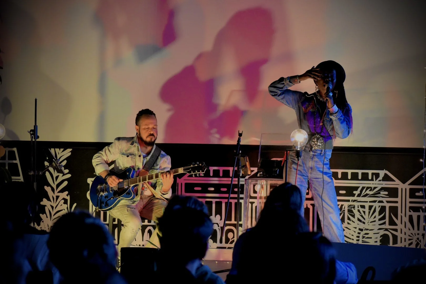 A live musical performance featuring a male guitarist seated and playing an electric guitar, and a female singer standing, holding a microphone, with her hand near her face. The stage has decorative elements and a large screen in the background proje