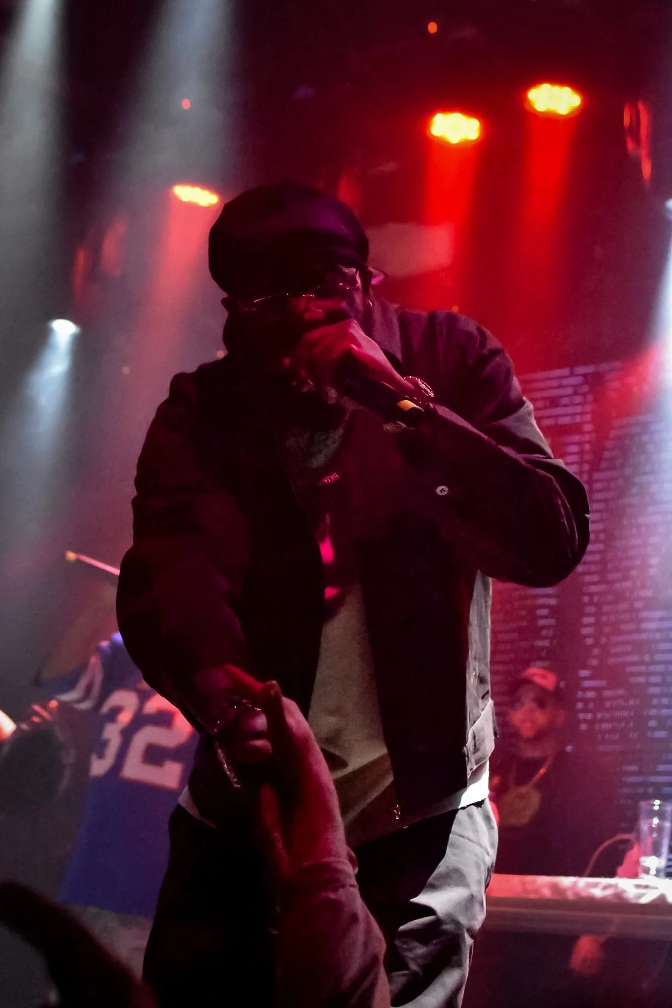 A man in a black beanie, glasses, and dark clothing is on stage in front of colorful lights, possibly at a concert or club. The background has red and white lighting effects, and there are other people visible in the crowd.