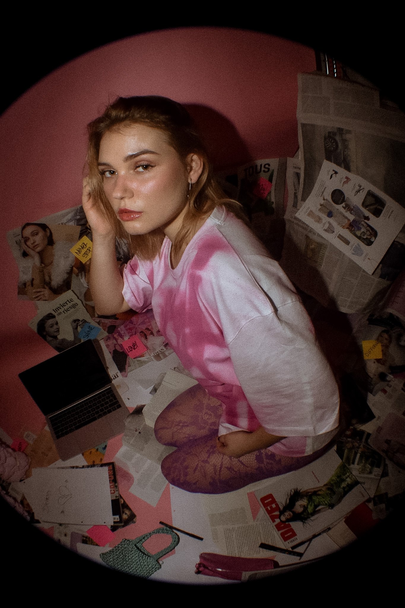 A young woman with shoulder-length blonde hair and makeup, wearing a pink and white tie-dye shirt and patterned tights, sitting on the floor of a cluttered room with magazines, papers, and a laptop.