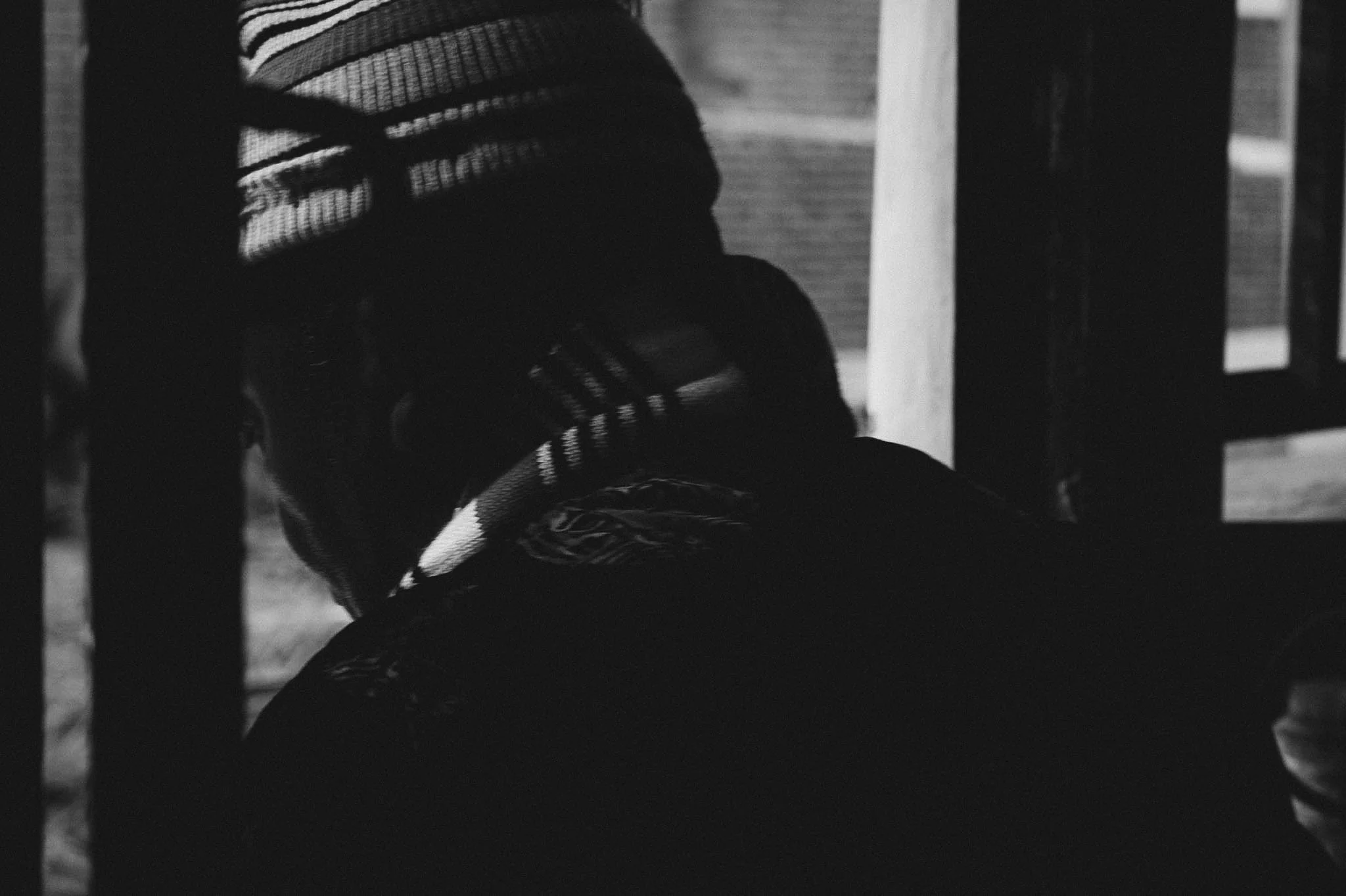 Black and white photo of a person wearing striped clothing, sitting on a bench with their head lowered, partially obscured.