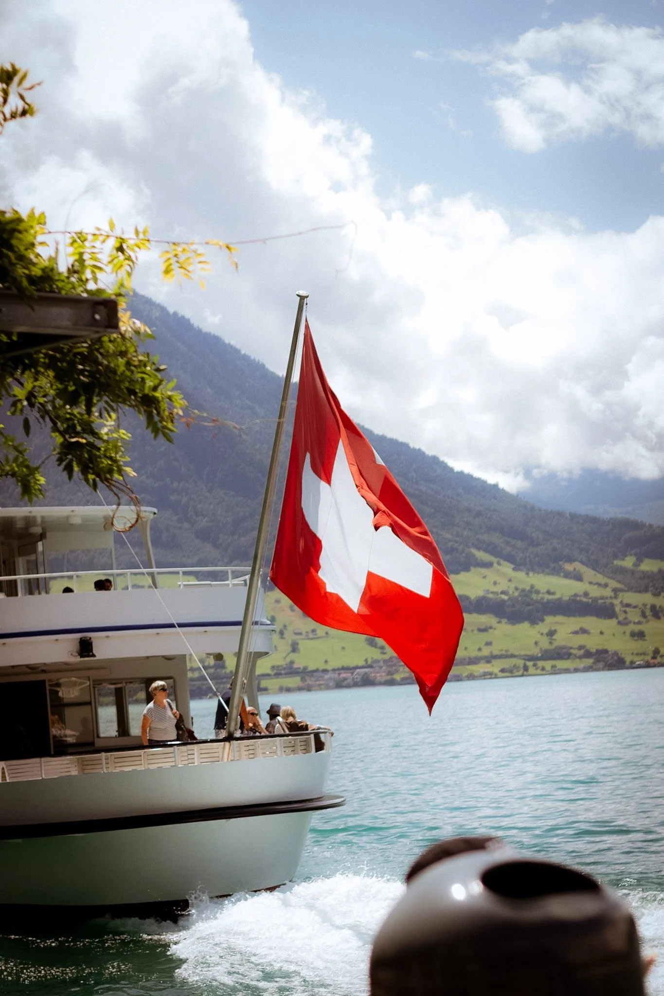 A boat with a red and white Swiss flag sailing on a lake, with mountains and a partly cloudy sky in the background.