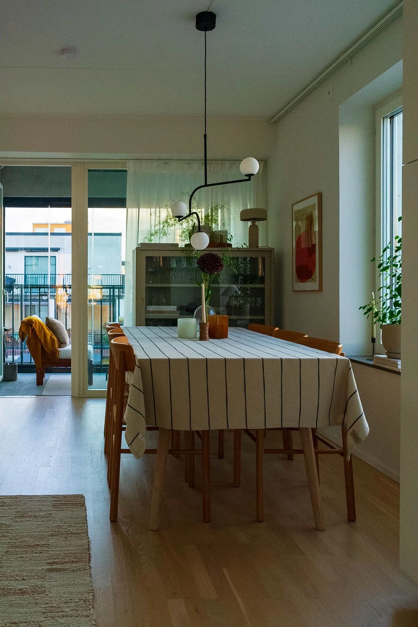 A dining room with a rectangular wooden table covered with a striped tablecloth, surrounded by six wooden chairs. There is a vase with a dark round flower on the table. The room has large windows with sheer curtains, letting in natural light. In the 