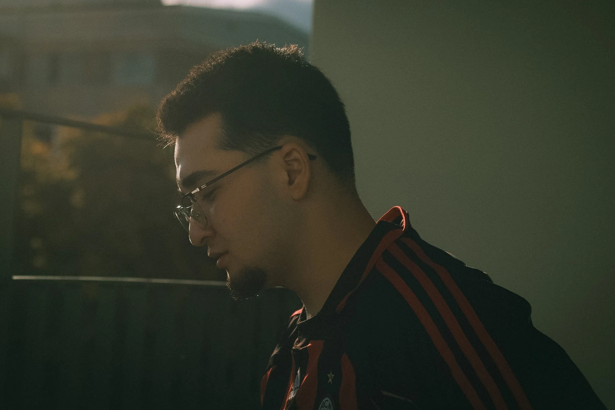 A young man with dark hair and glasses looking down, wearing a black and red striped jacket with a logo on it, with a dark background and soft lighting.