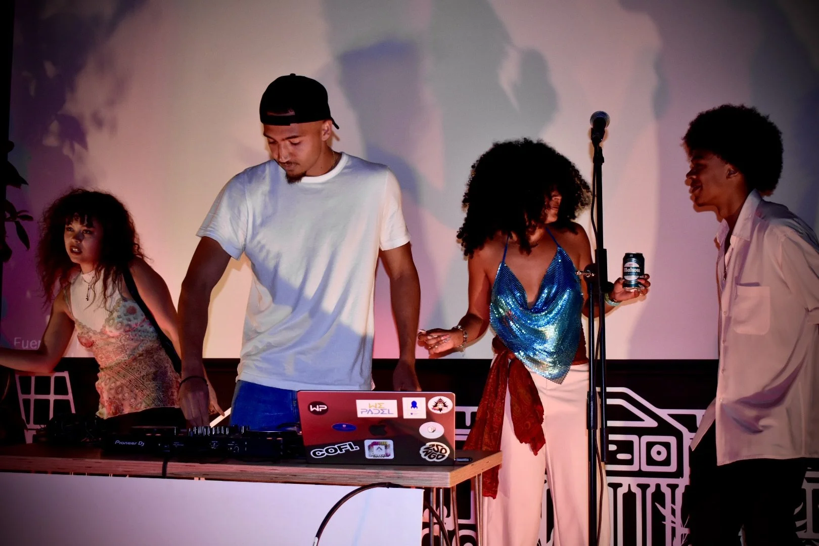 Group of young adults at a party with a DJ, some holding drinks, in a dimly lit indoor space.