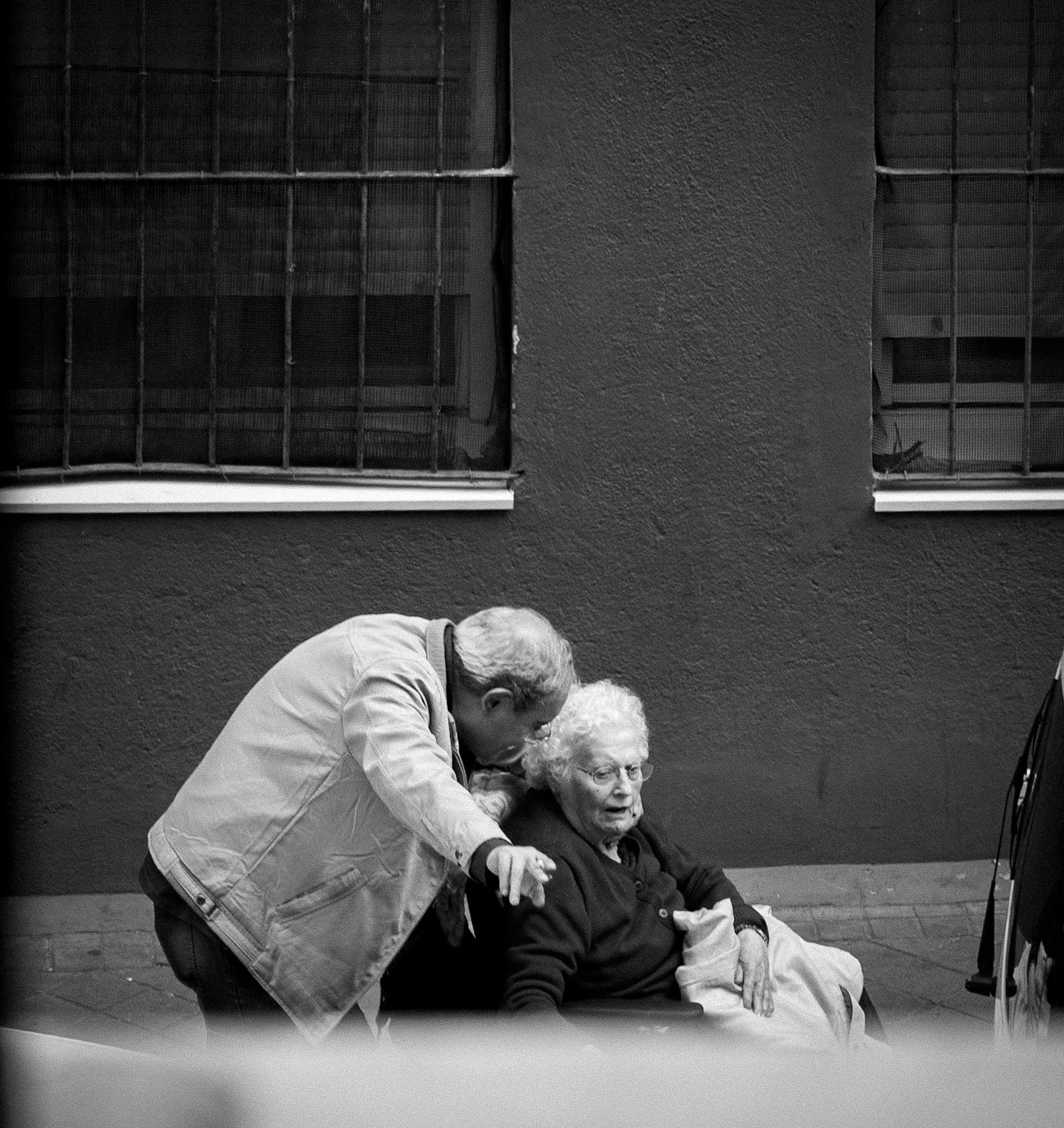 Black and white photo of a person with white hair and glasses leaning over and comforting another older person sitting on a bench outside, along with a dog. There are barred windows on a brick wall behind them.