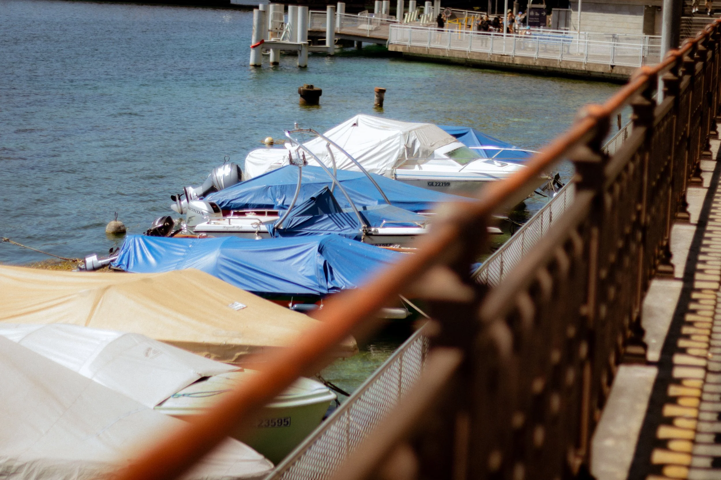 Boats docked by the water with blue covers and outboard motors, viewed through a chain-link fence and a wooden walkway.