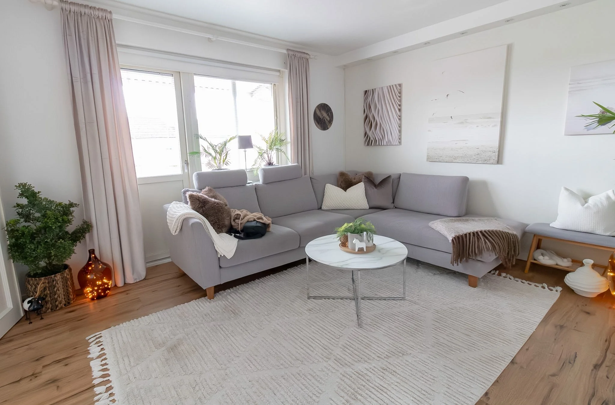 Living room with a large gray sectional sofa, white wall with art, plants near window, round white coffee table, and beige area rug.