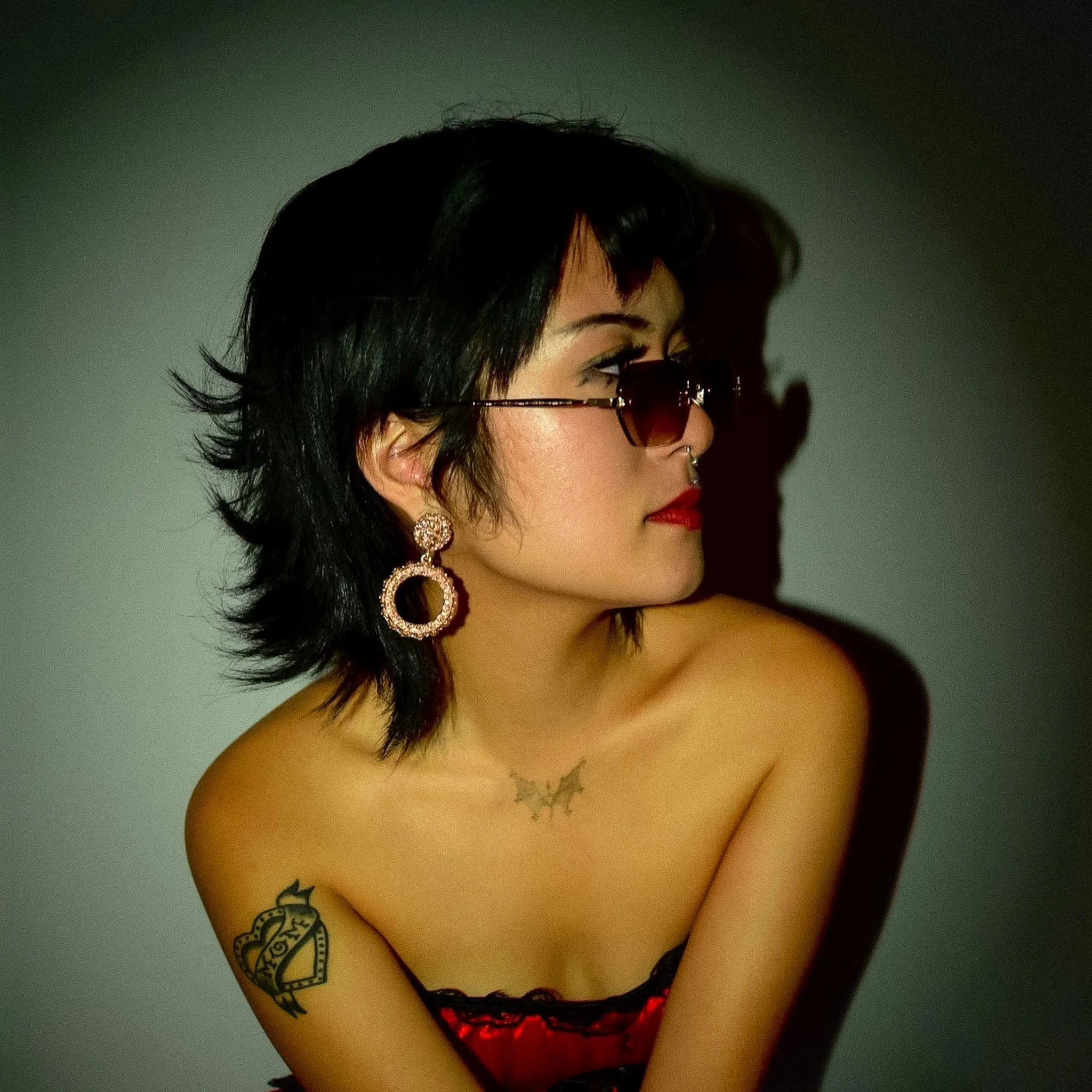 Close-up of a woman with black hair, wearing sunglasses, red lipstick, and large circular earrings. She is topless, showing tattoos on her shoulder and chest, against a plain background.