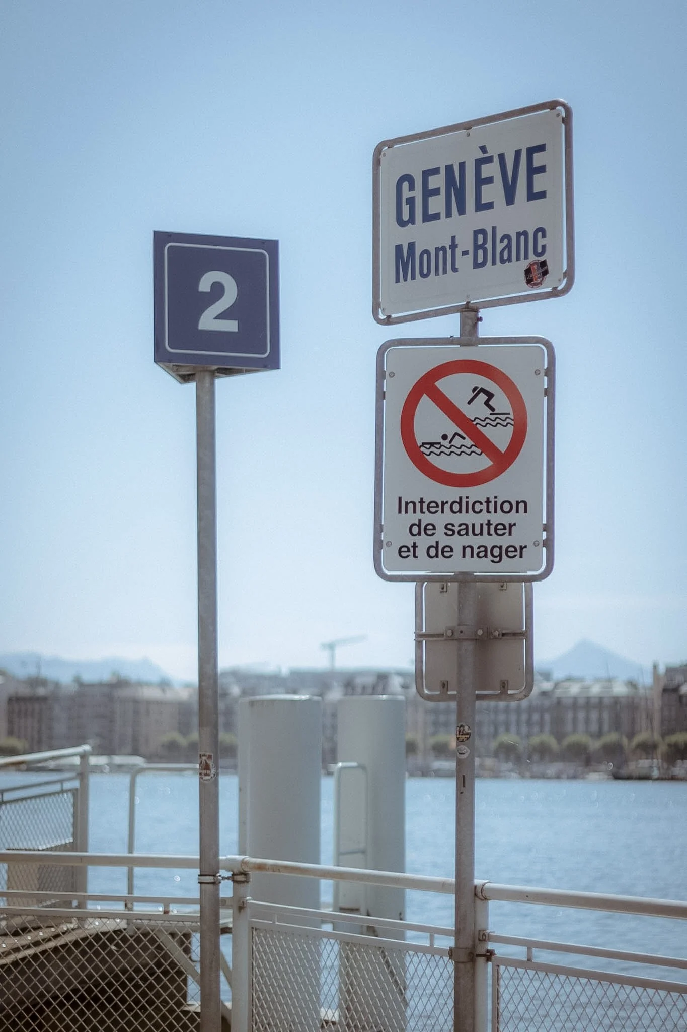 Signage on a boat indicating Geneva and Mont-Blanc, with a no swimming or jumping sign below, overlooking a body of water and a cityscape in the background.