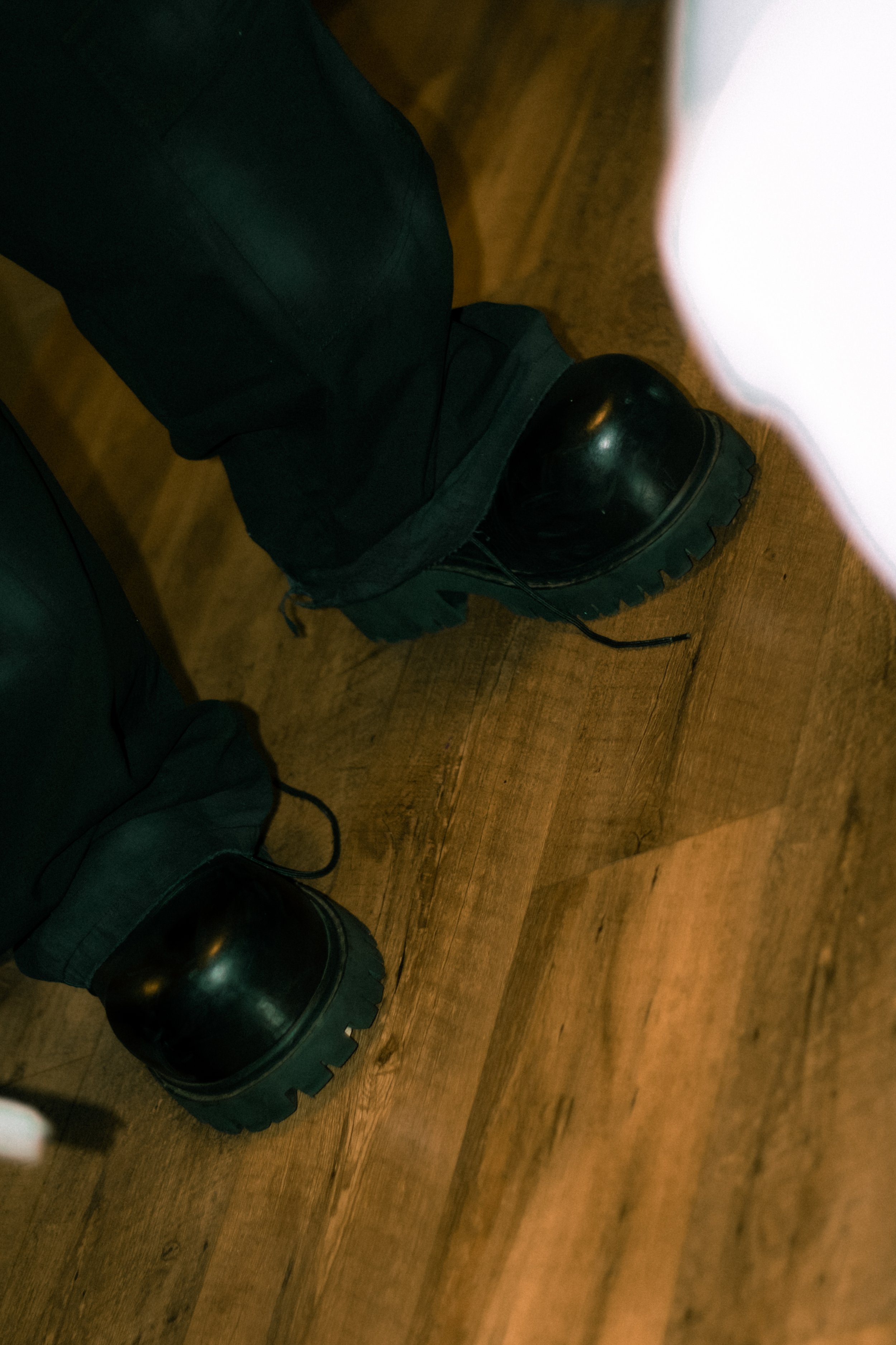 Close-up of a person wearing black leather boots and black pants, sitting on a wooden floor, with another person also sitting nearby; part of a white object is visible on the right side of the image.