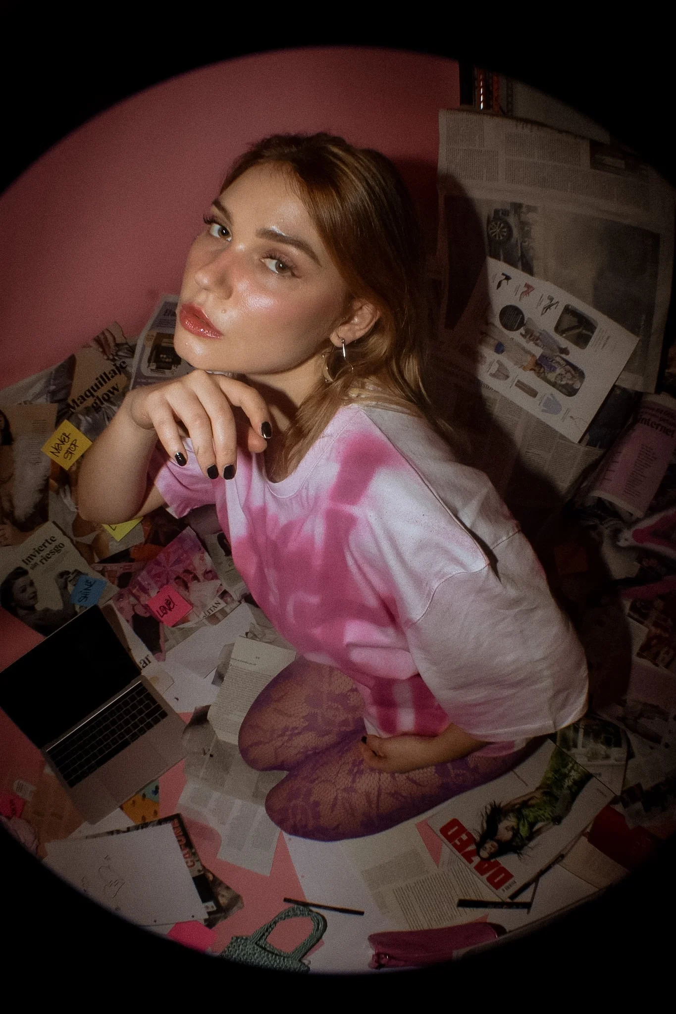 A young woman with brown hair, makeup, and hoop earrings posing in front of a pink wall, sitting on the floor with magazines around her, wearing a pink tie-dye shirt and purple fishnet stockings.