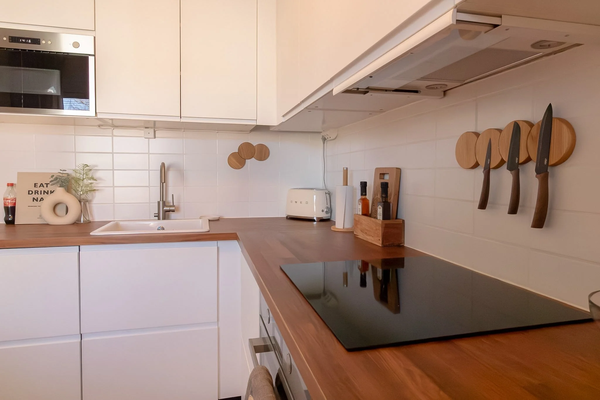 A modern kitchen with white cabinets, a wooden countertop, and a black electric stovetop. There is a white sink with a silver faucet, a toaster, and various kitchen utensils and decorations, including knives on wooden holders and a small plant. A mic