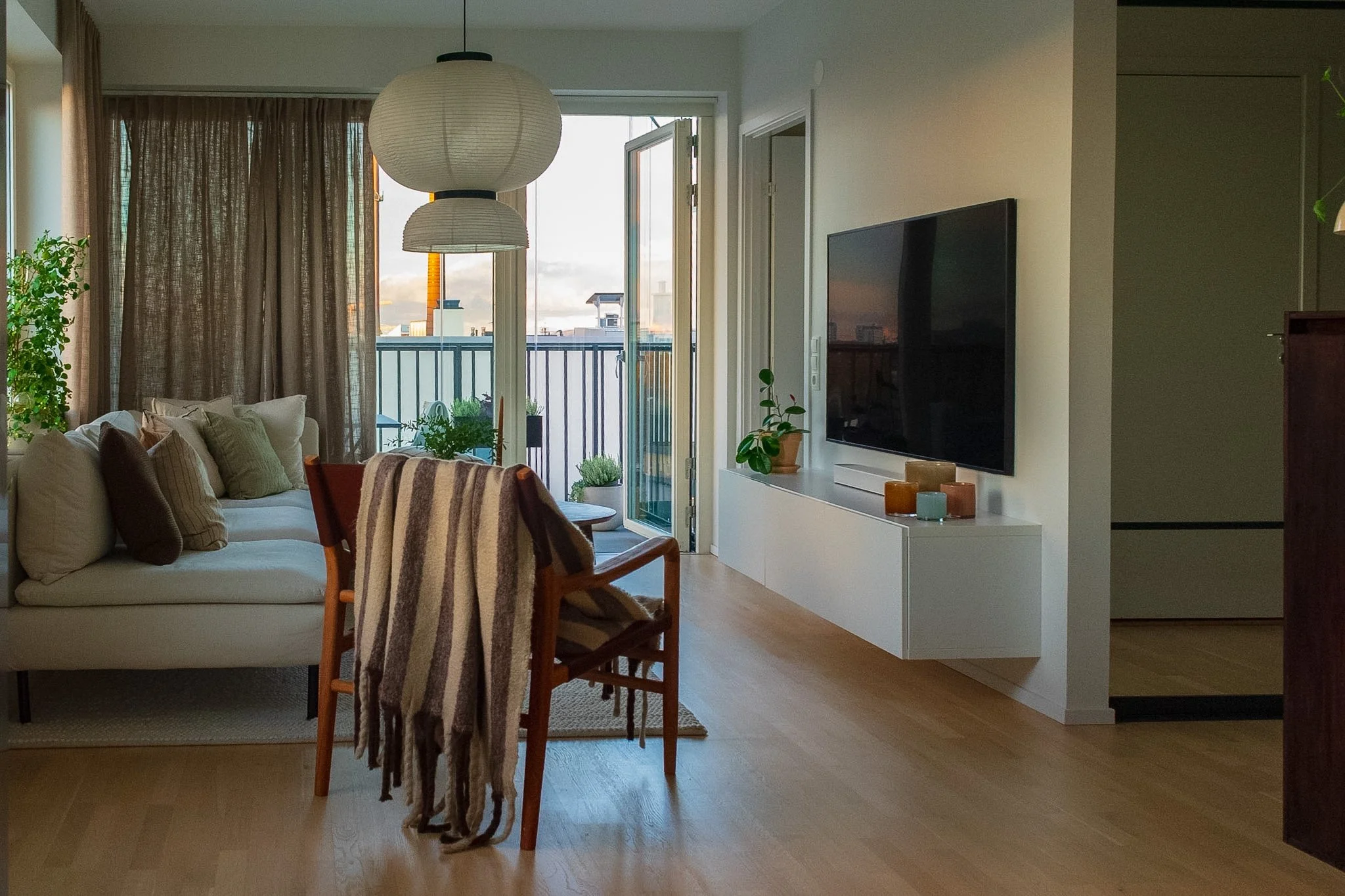Living room with beige couch, multiple pillows, wooden chairs with striped throw, large window with curtains, balcony door open showing an outdoor cityscape, television on white stand, potted plant, pendant light hanging from ceiling.