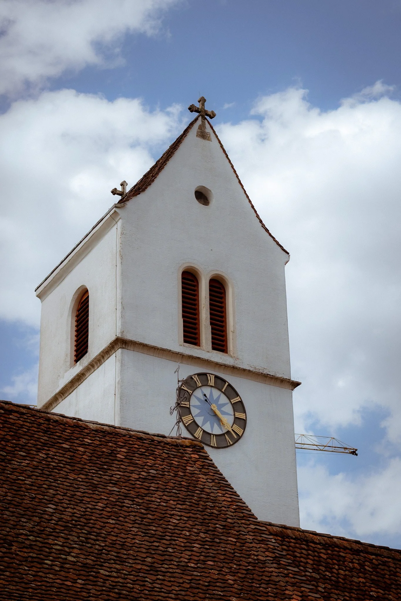 A white church tower with a clock showing almost 11:30, under a partly cloudy sky. The church has small arched windows with wooden shutters and a steep, reddish-tiled roof in the foreground.