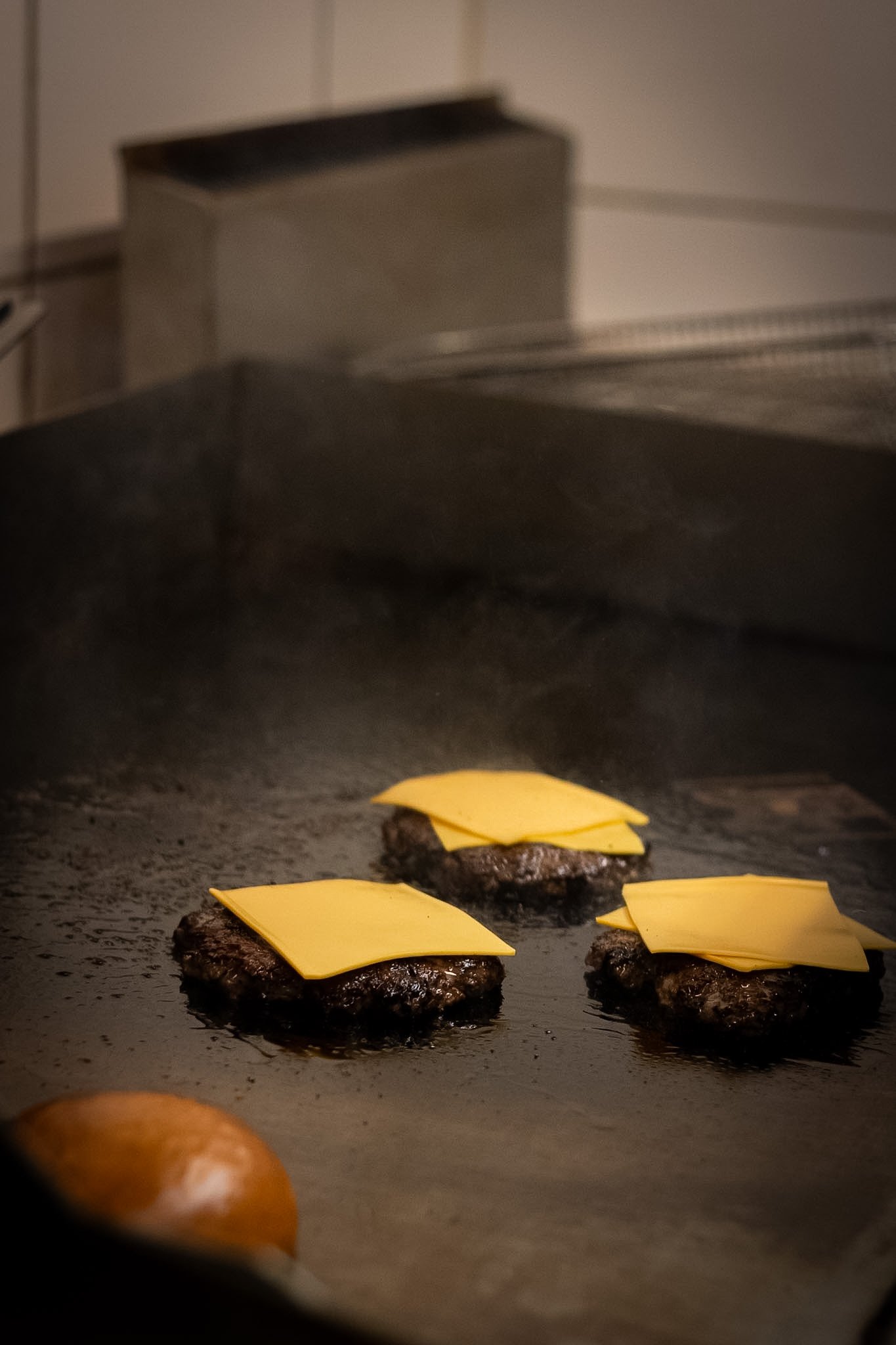 Close-up of three raw burger patties topped with slices of cheddar cheese on a baking sheet, with a blurred oven in the background.