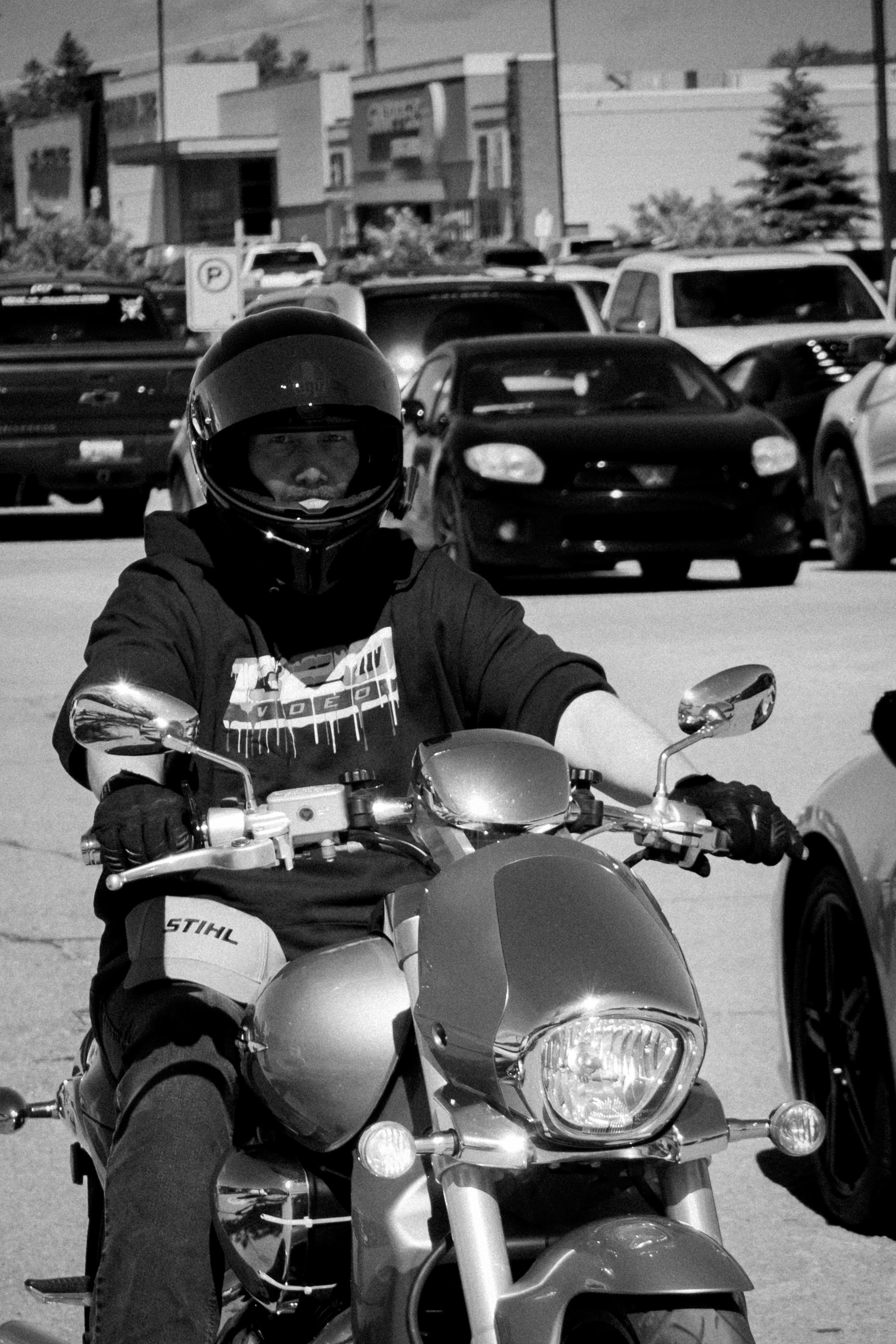 A person wearing a helmet and riding a motorcycle in a parking lot with multiple parked cars and buildings in the background.