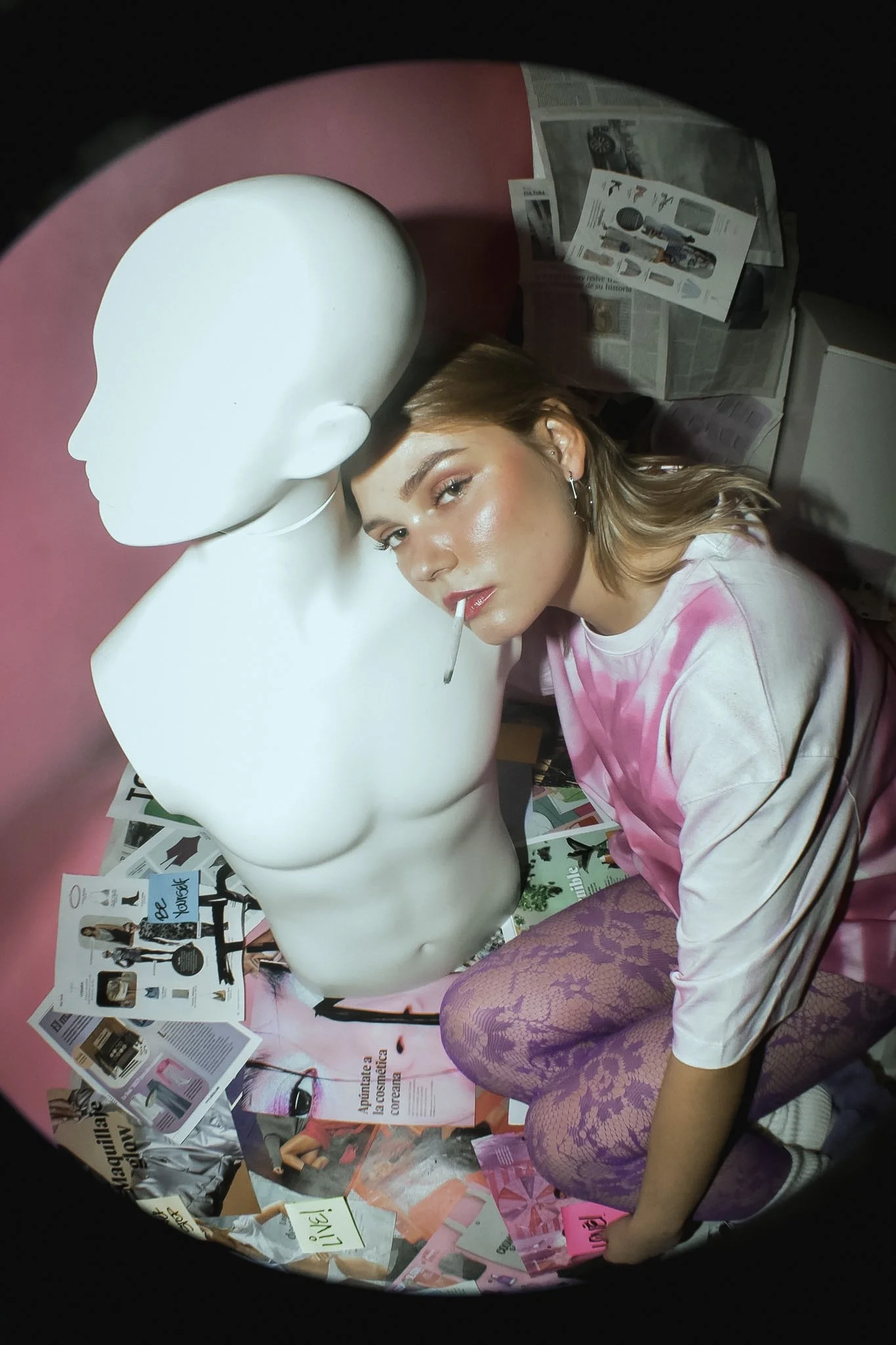 A woman with light skin and blonde hair wearing a pink tie-dye sweatshirt and purple lace stockings is crouching next to a white mannequin head. The woman is holding a pink item and has a lollipop in her mouth. The background has magazines, pamphlets