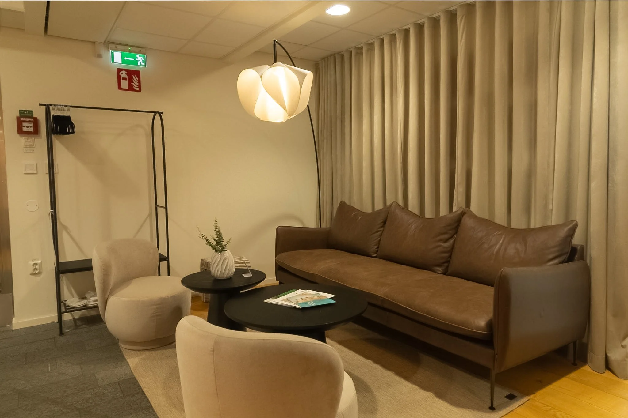 A cozy room with a brown leather sofa, cream-colored armchairs, a small round black table with magazines, a decorative vase with greenery, and a contemporary white pendant lamp, all against cream-colored curtains.