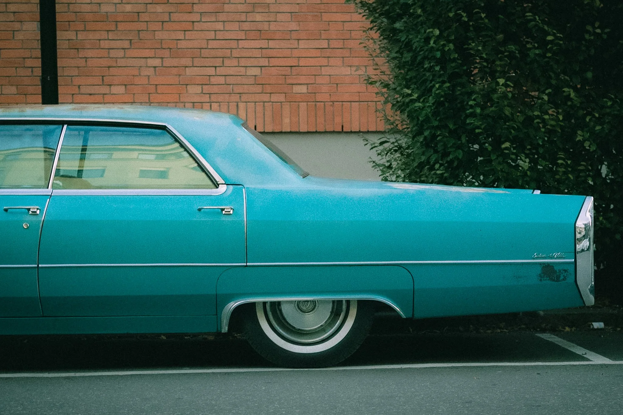 Side view of a vintage blue car parked next to a brick wall with a green bush nearby.