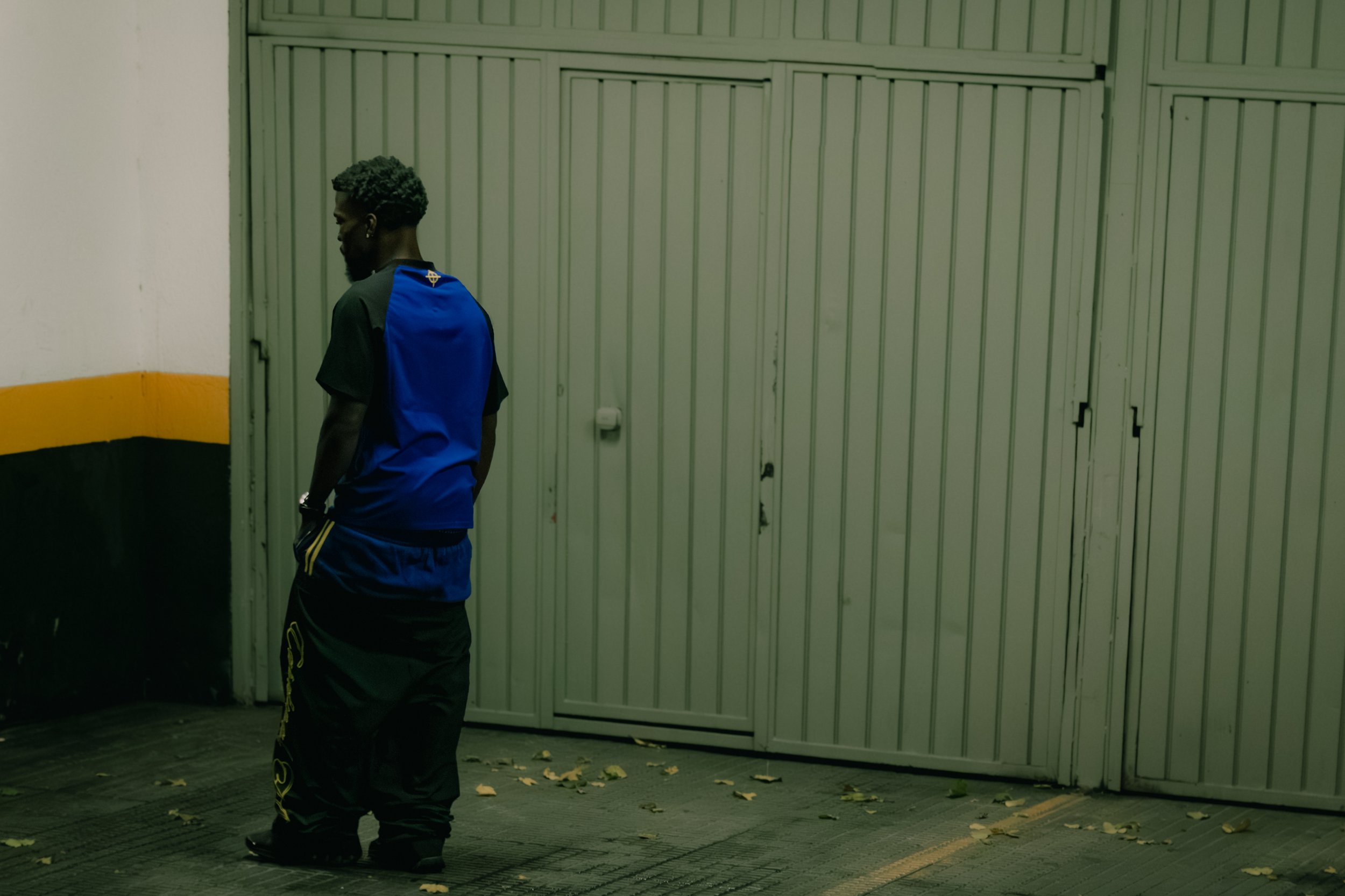 A man wearing a blue and black sports shirt and black pants stands on a sidewalk next to a large, closed metal gate. The gate is light green, and the surrounding wall has white, yellow, and black sections. Fallen leaves are scattered on the ground.