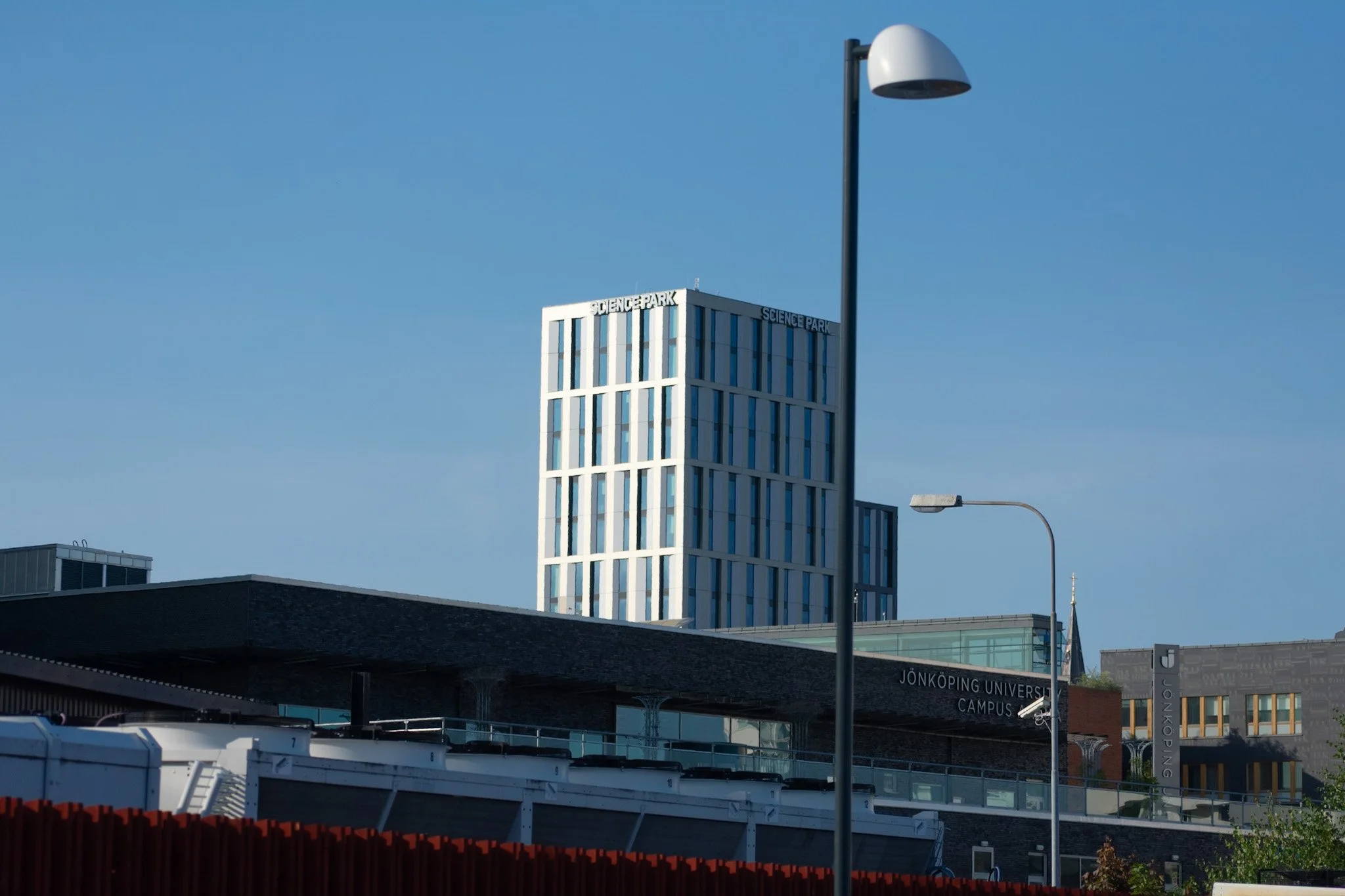A tall building labeled 'Science Park' with modern architecture and blue sky background. In the foreground, a street lamp, some vehicles, and a sign for Jonkoping University Campus are visible.