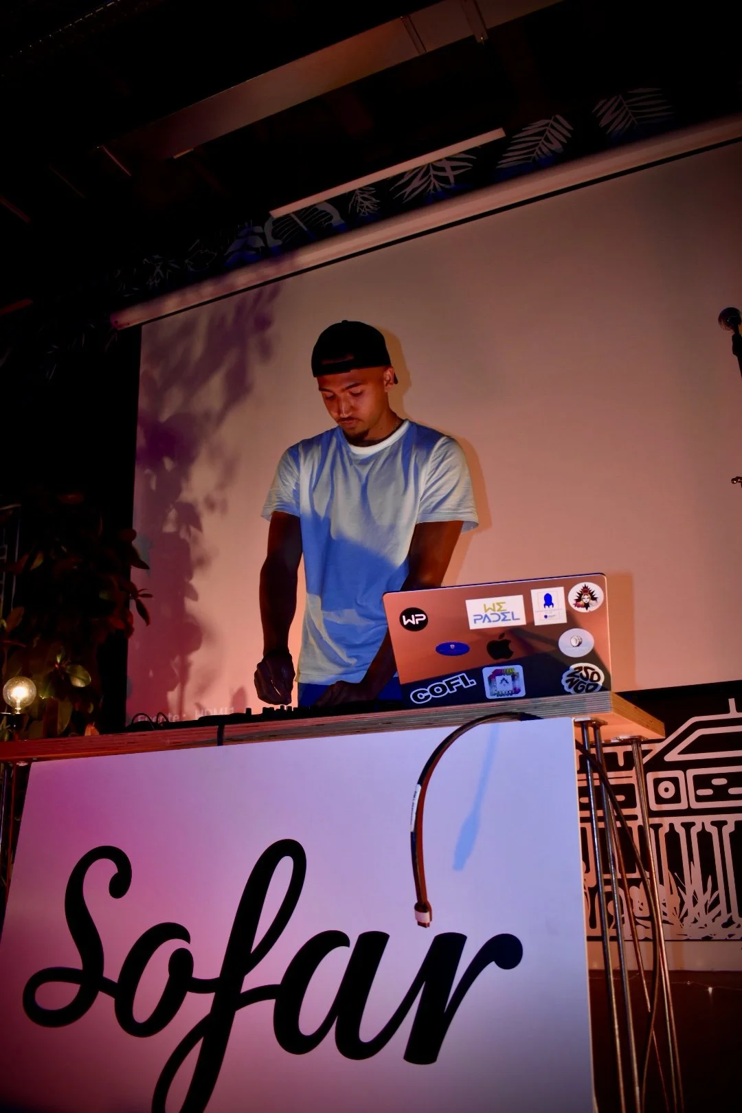 A young man wearing a black cap and white t-shirt, standing behind a DJ booth with a laptop covered in stickers, performing at an event with a decorated backdrop. The booth has a large sign with the word 'Sofyar' on it.