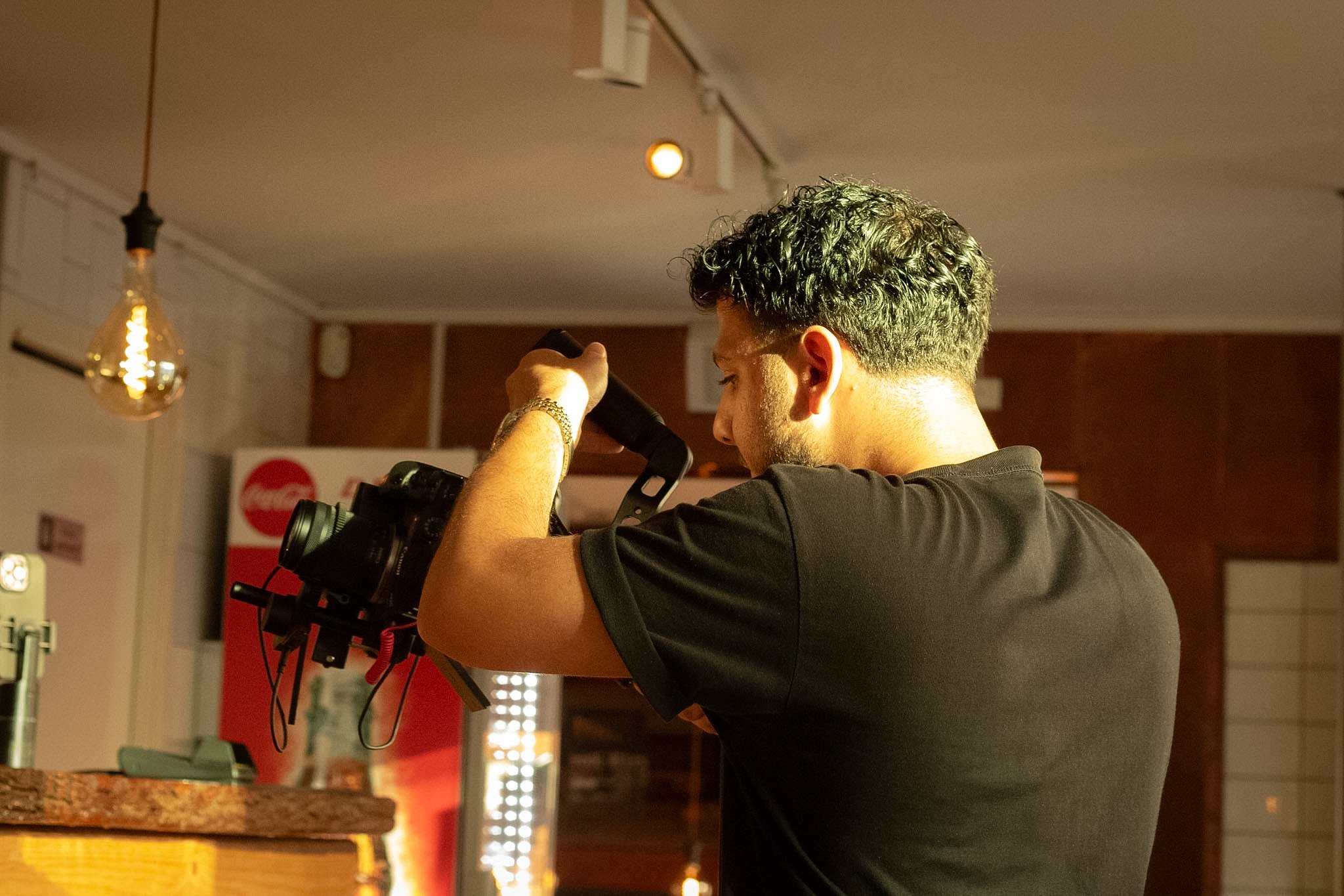 A man with curly hair operating a camera inside a building with warm lighting and pendant lights hanging from the ceiling.