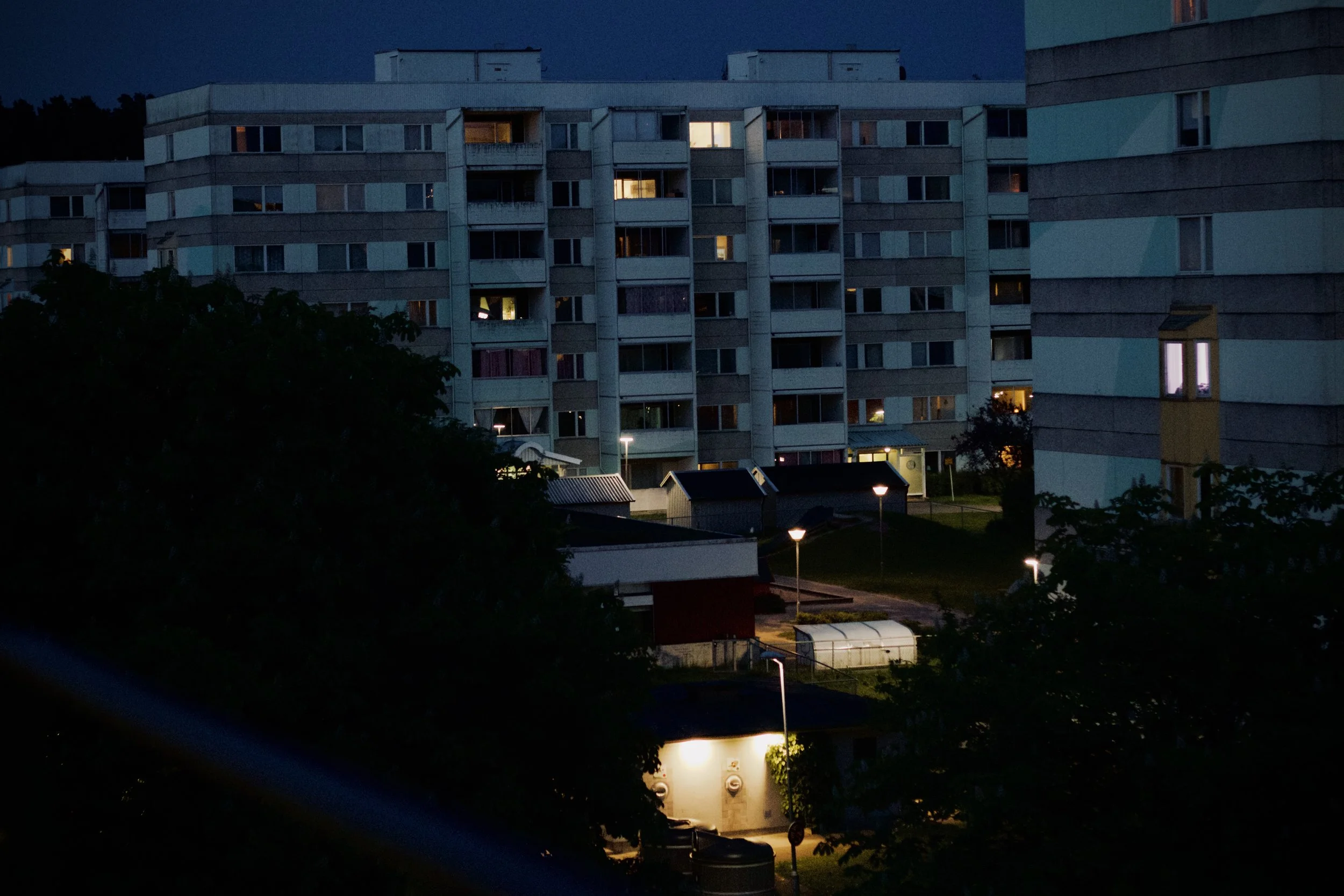 Nighttime view of a multi-story apartment building with some windows illuminated, and streetlights and nearby structures visible in the foreground.