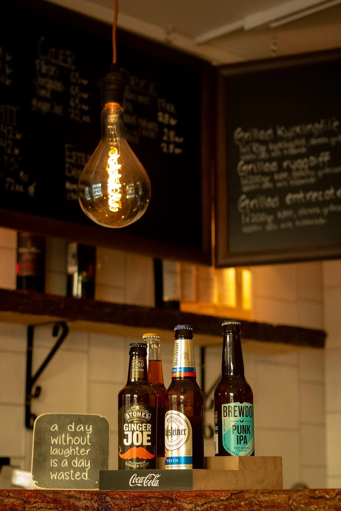 A row of three bottled drinks with a blackboard menu and a motivational quote in the background, illuminated by a hanging light bulb.