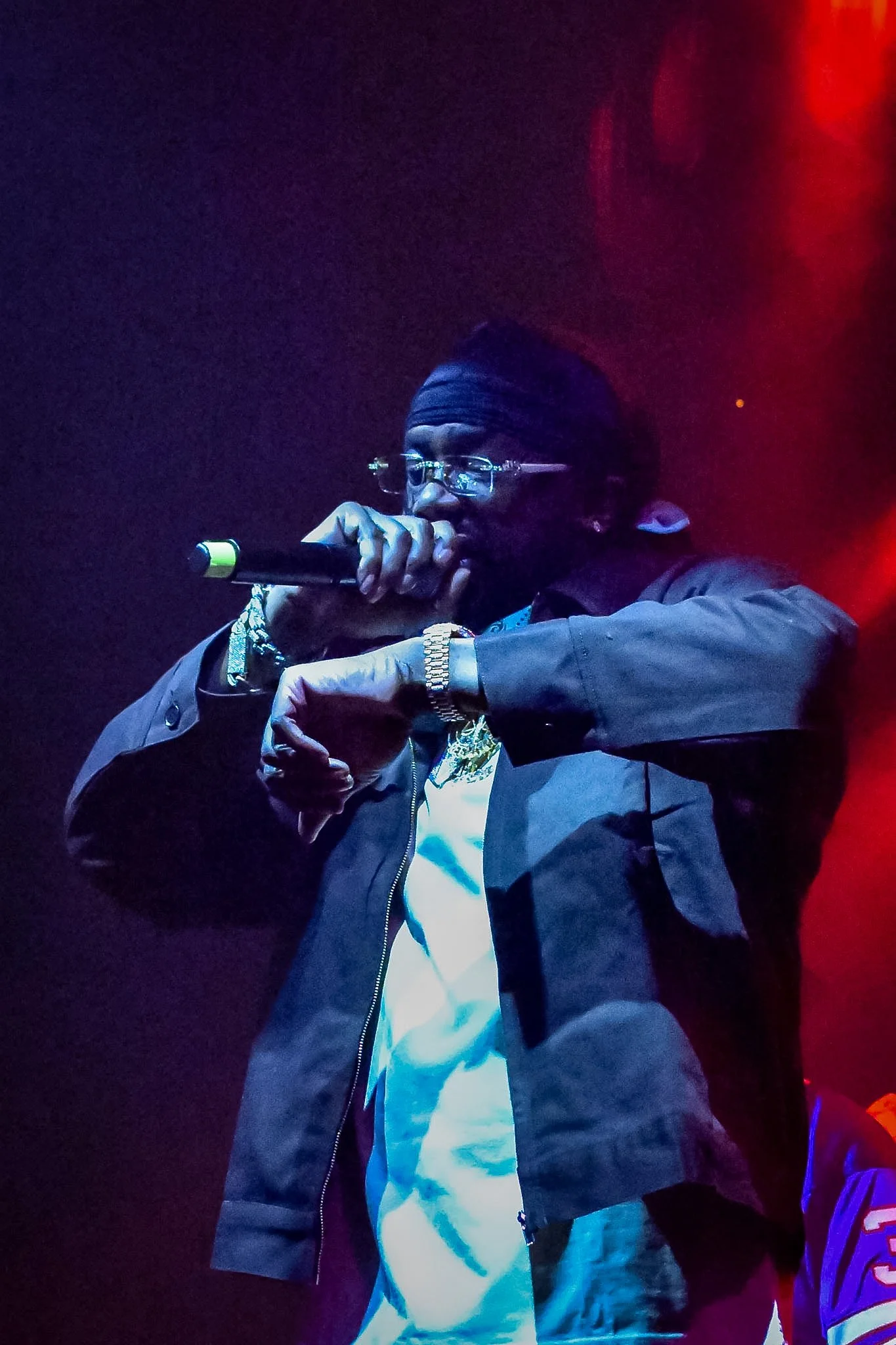A person wearing glasses, a black beanie, and a dark jacket singing or speaking into a microphone on stage, with colorful stage lighting around.