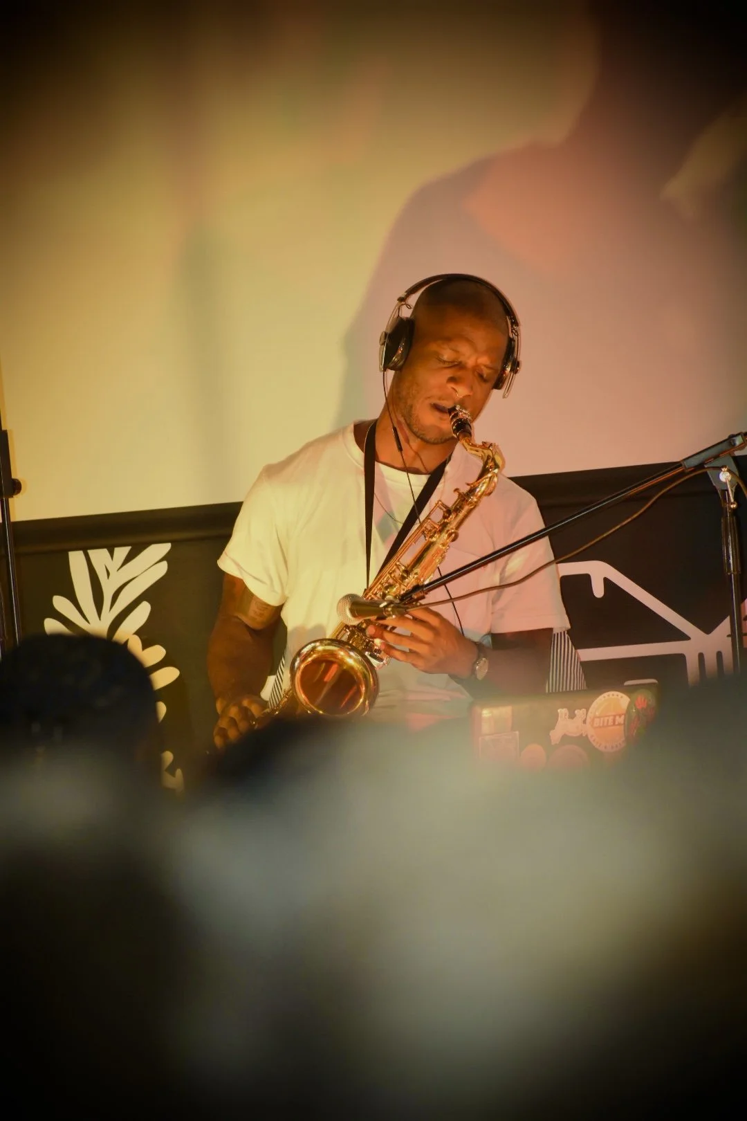 A man in a white t-shirt playing a saxophone during a performance, wearing headphones, on a stage with a backdrop that has a black and white design.