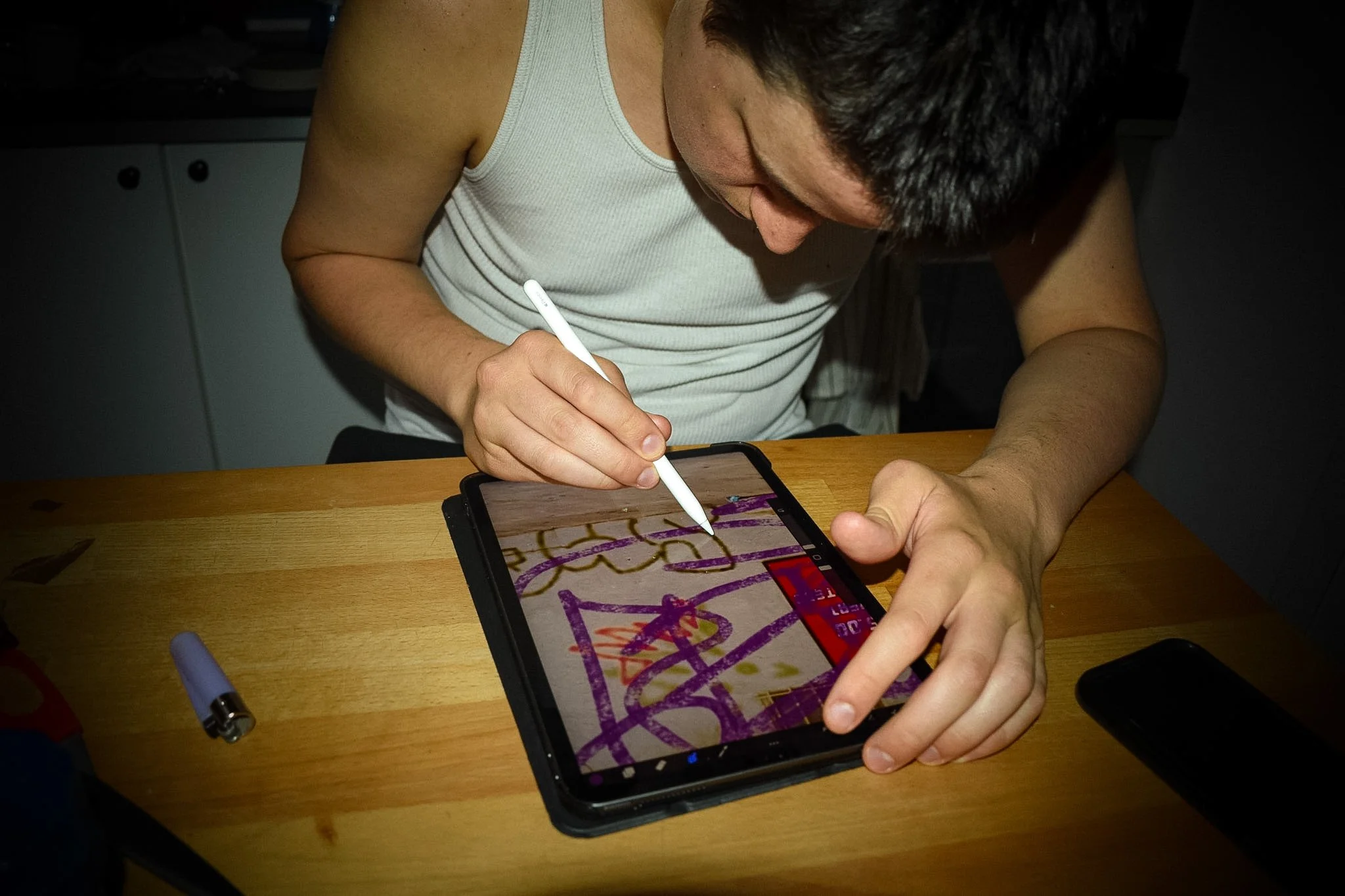 A person using a stylus to draw or write on a tablet screen, with a colorful drawing visible on the screen. The tablet is placed on a wooden table with a gray marker and a black object nearby.