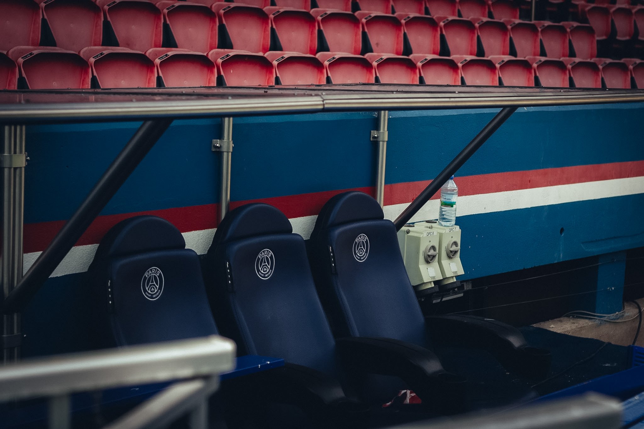 Empty red stadium seats, black cushioned seats with Paris Saint-Germain logo, water bottle, and electrical outlets on the blue wall.